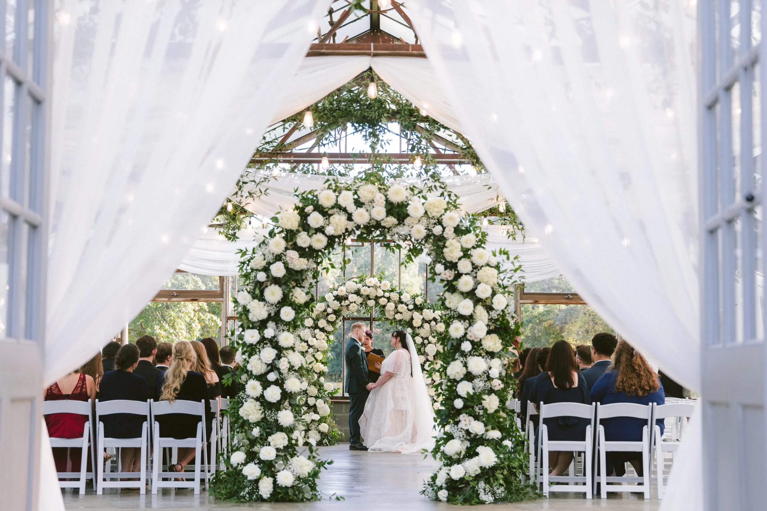 Outdoor wedding or event reception setup with a long wooden table decorated with floral arrangements, candles, and string lights hanging above, surrounded by wooden chairs and lush green trees in the background.