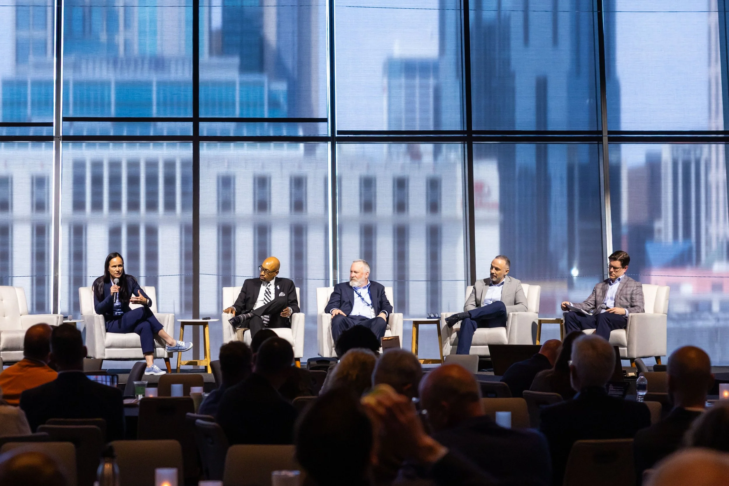 A panel of five people sitting on stage during a conference or panel discussion, with a large window and cityscape in the background, and audience members seated in front.