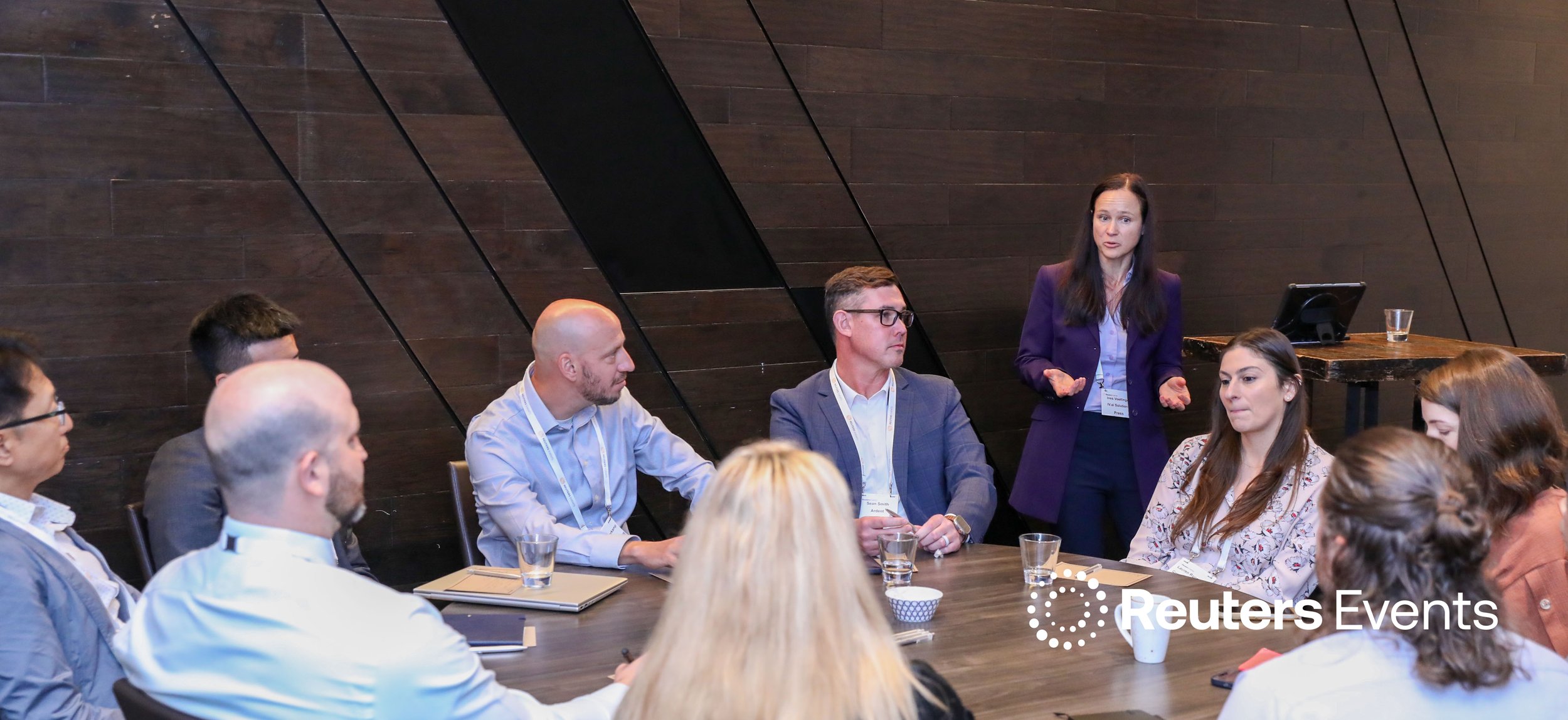 A woman in a purple blazer speaking to a seated group of diverse adults at a conference table in a modern room with dark wooden walls. A logo for Reuters Events is visible in the bottom right corner.