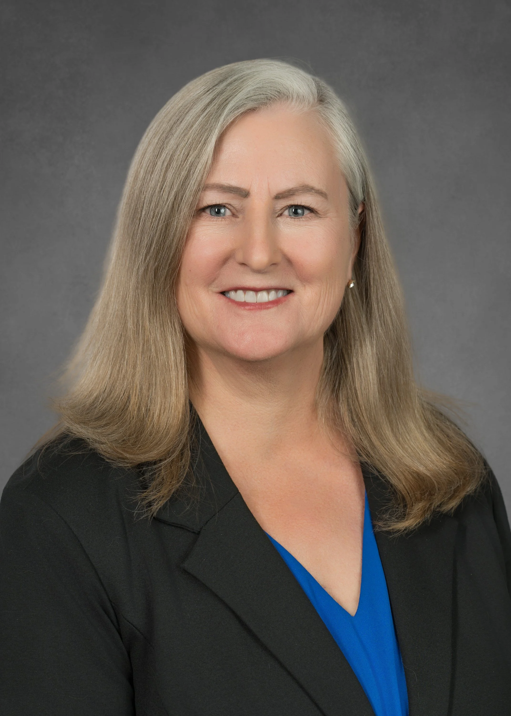 Portrait of a middle-aged woman with long blonde hair, blue eyes, and a friendly smile, wearing a black blazer over a blue blouse, against a gray background.