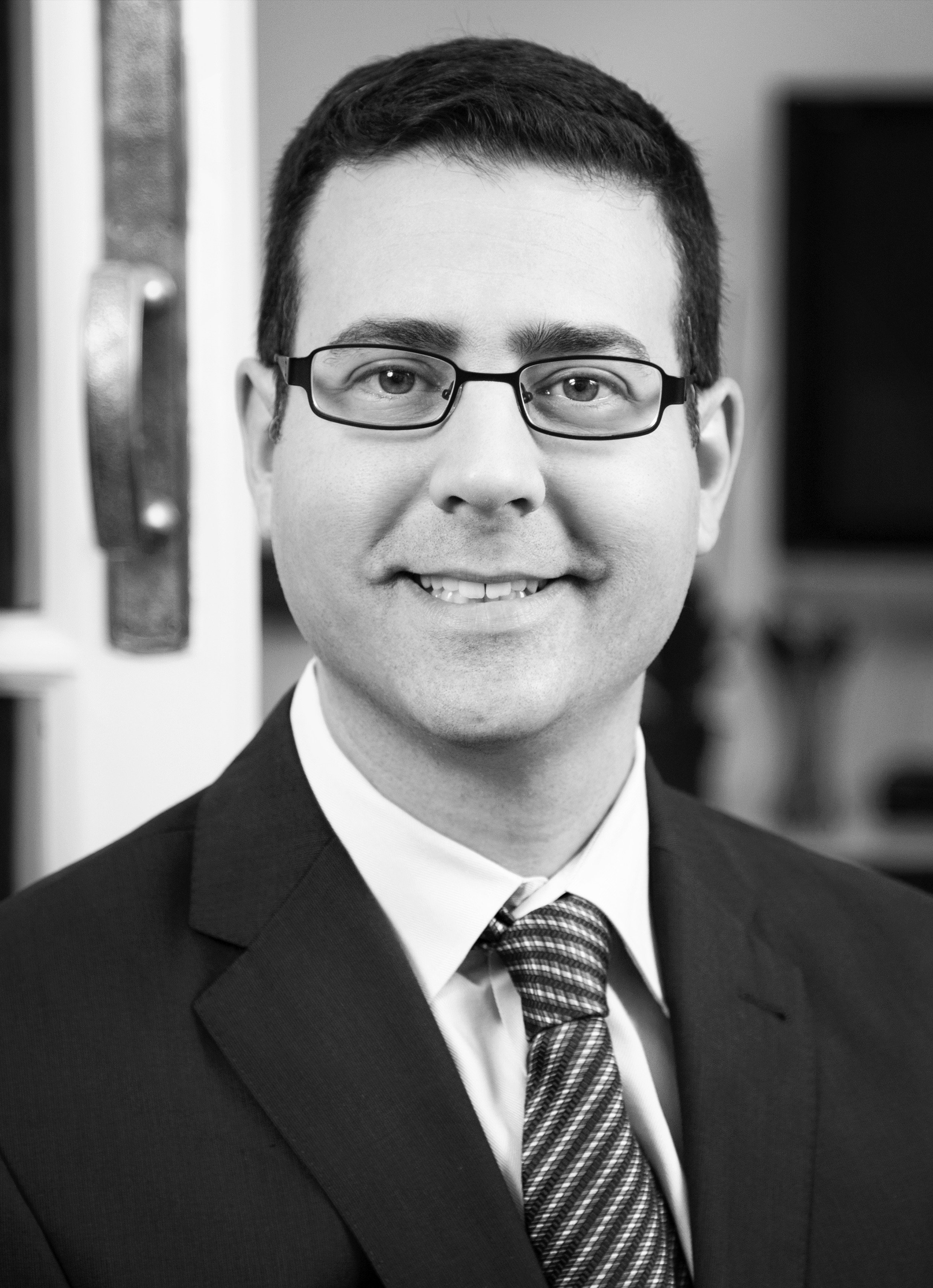 Black and white photo of a man in a suit, wearing glasses, smiling, with a striped tie, out of focus background.