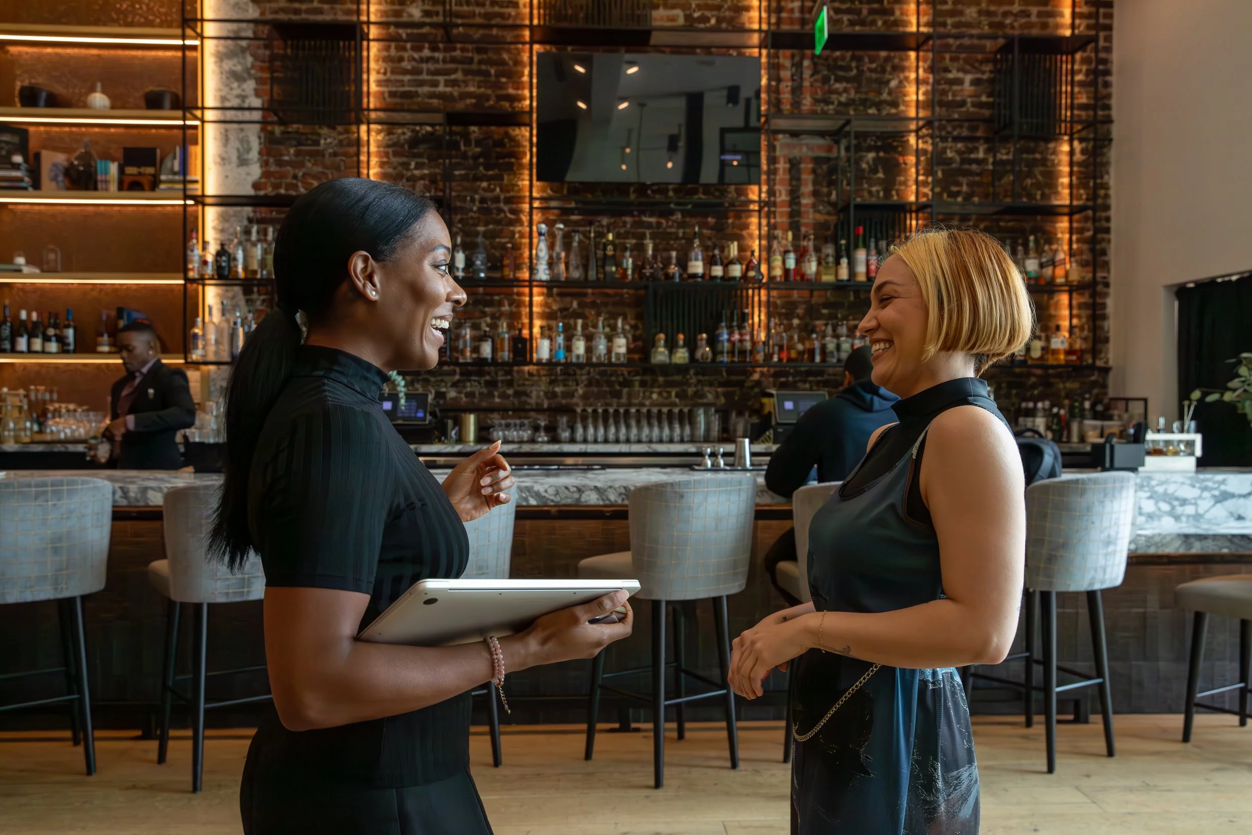 Two women are smiling and talking in a bar or restaurant setting. One woman has black hair and is holding a tablet, while the other has short blonde hair and is wearing a sleeveless top. The background shows shelves with bottles and bar equipment, with a brick wall and a TV screen.