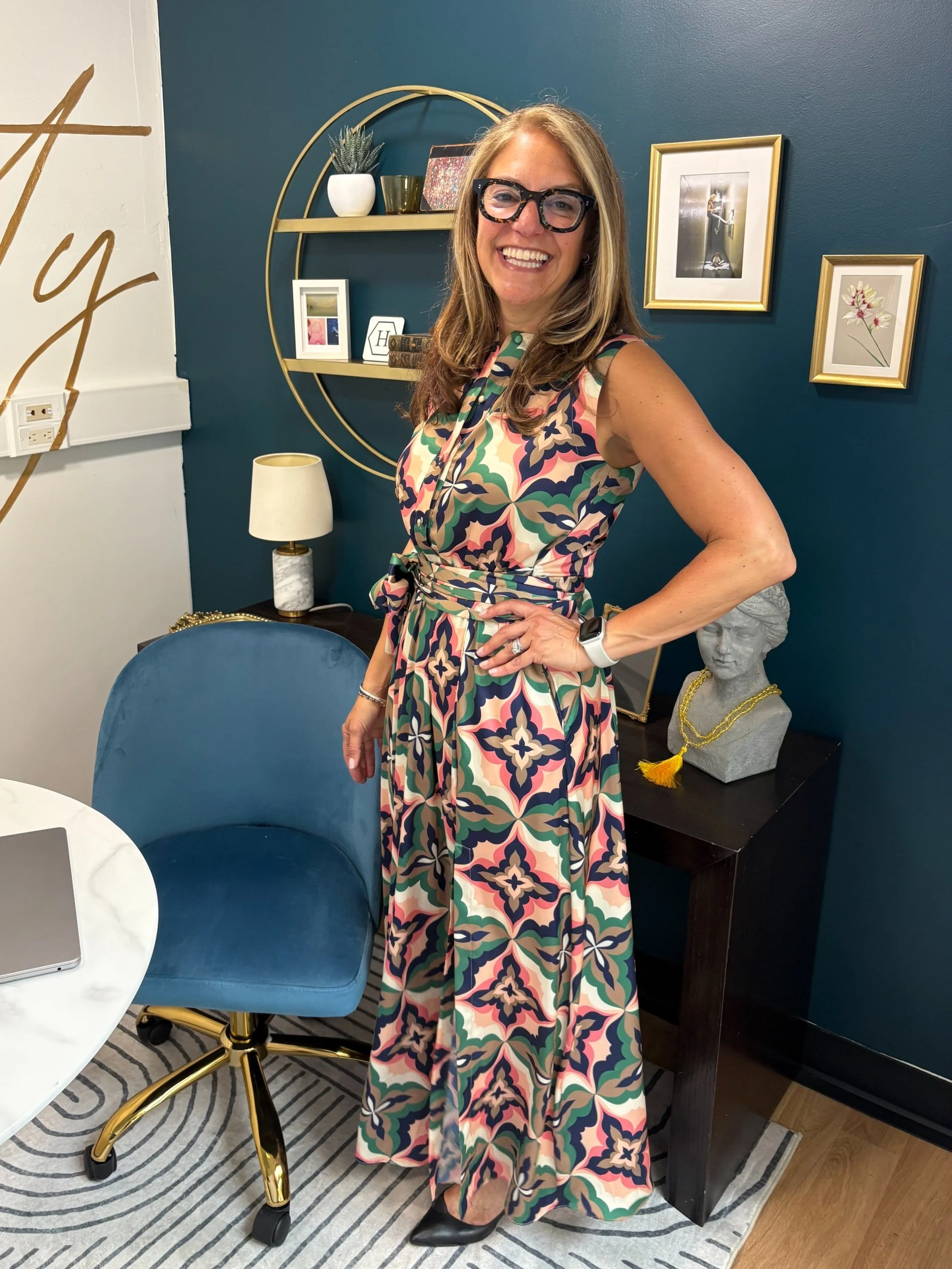 A woman with long hair, wearing glasses and a patterned sleeveless dress, standing in an office next to a table with decorative items and framed pictures on the wall behind her.