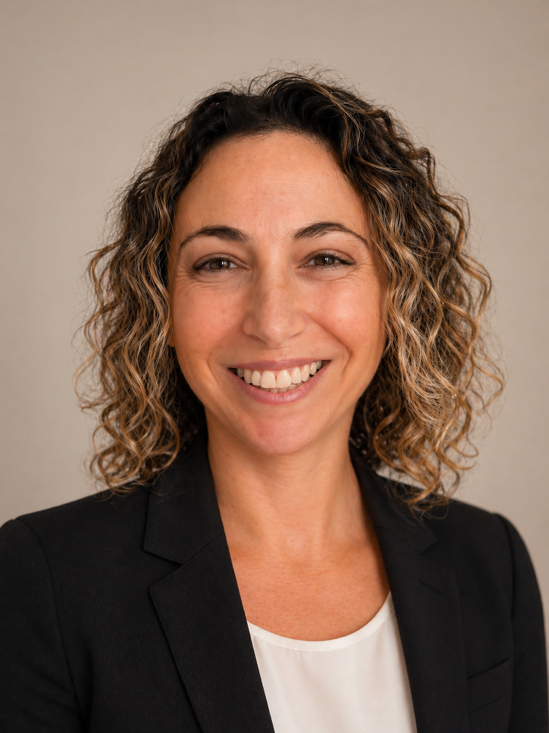 A woman with curly brown hair smiling, wearing a black blazer over a white top.