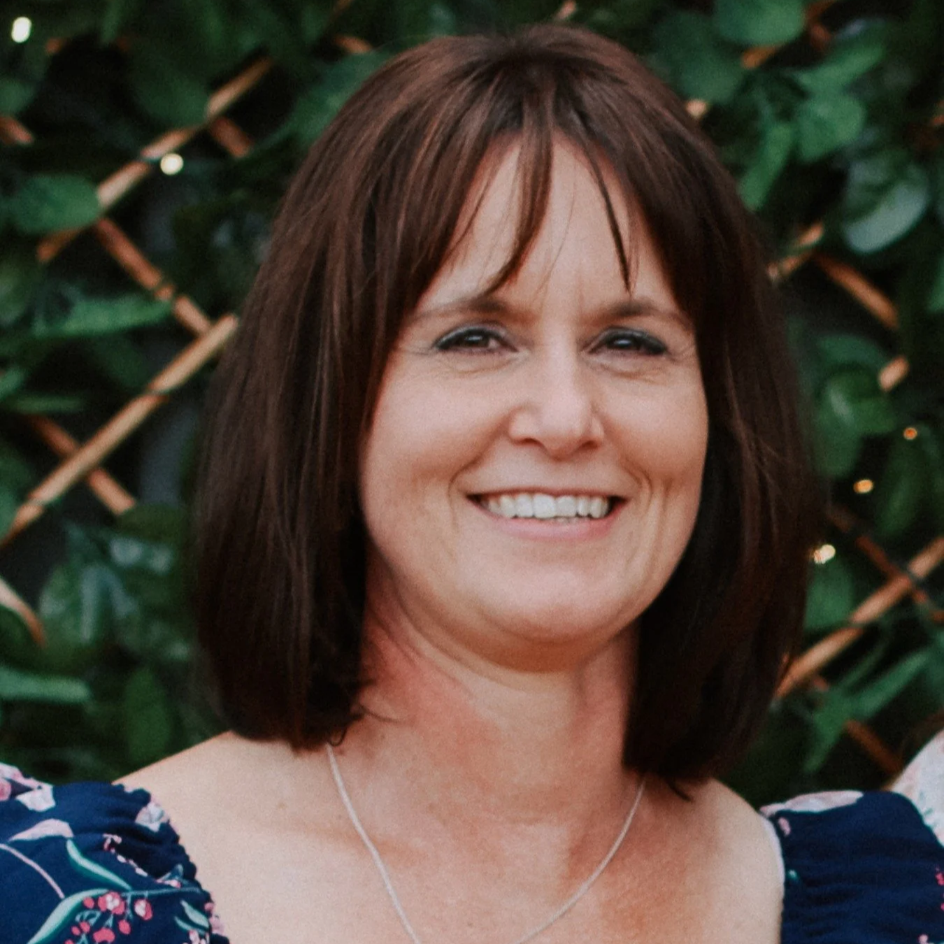 Black and white photo of a woman with shoulder-length dark hair, smiling, wearing a floral dress and a necklace, standing in front of a background with foliage and wooden lattice.