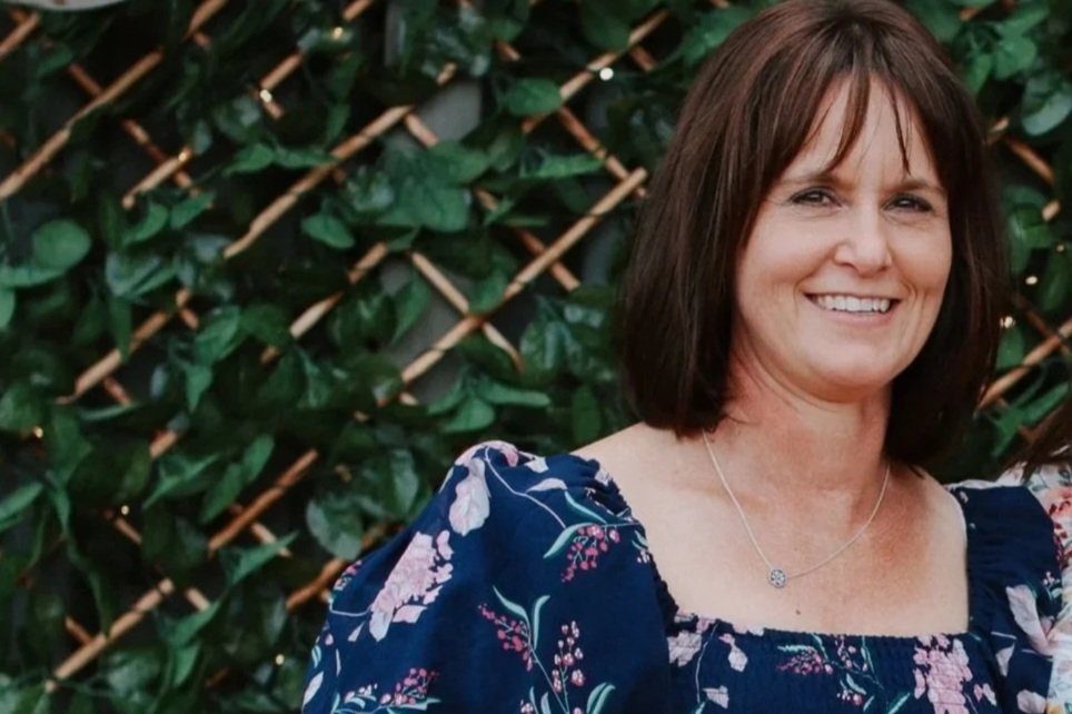 Black and white photo of a woman with shoulder-length dark hair, smiling, wearing a floral dress and a necklace, standing in front of a background with foliage and wooden lattice.