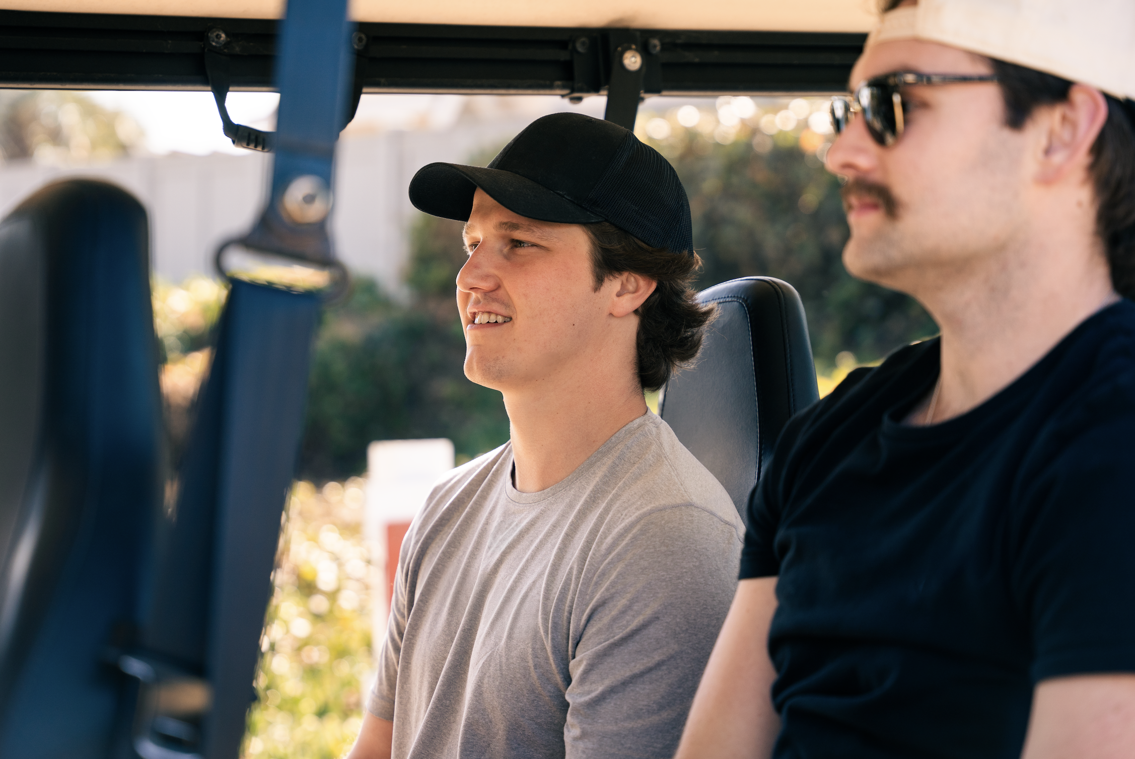 Two men sitting in a golf cart outdoors during daytime, one wearing a black cap and gray t-shirt, the other wearing a beige cap, sunglasses, and black t-shirt, with trees and a fence in the background.
