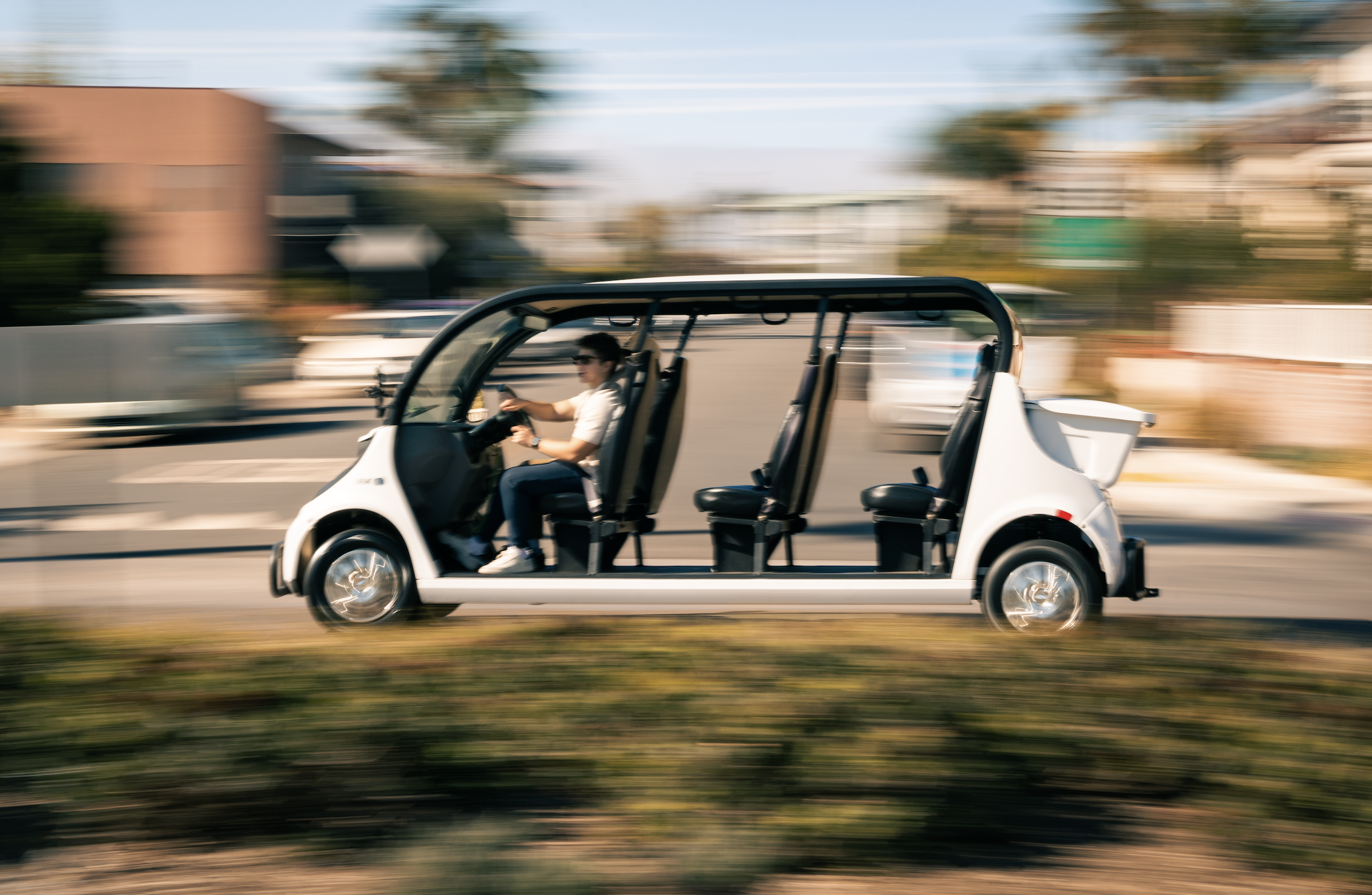 A person driving a white multi-seated electric vehicle on a city street with motion blur in the background.