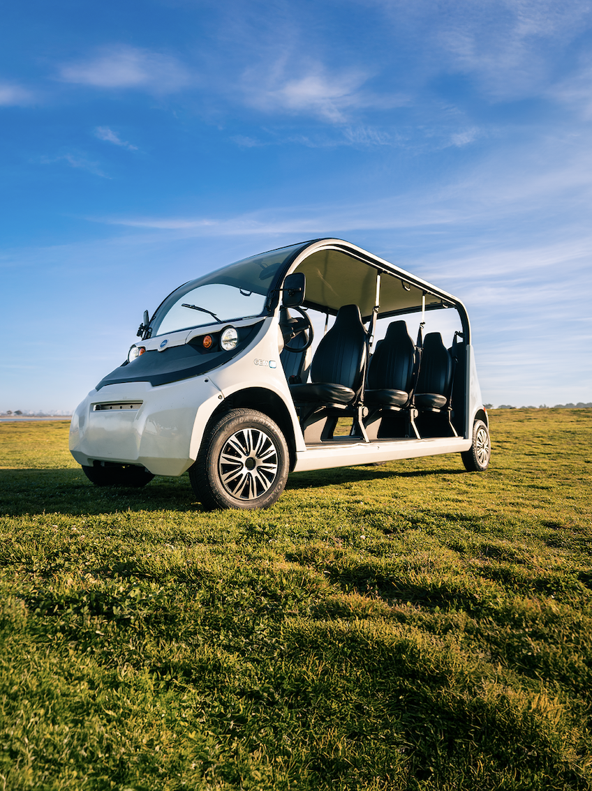 A small electric shuttle vehicle with four seats, parked on grass under a blue sky.
