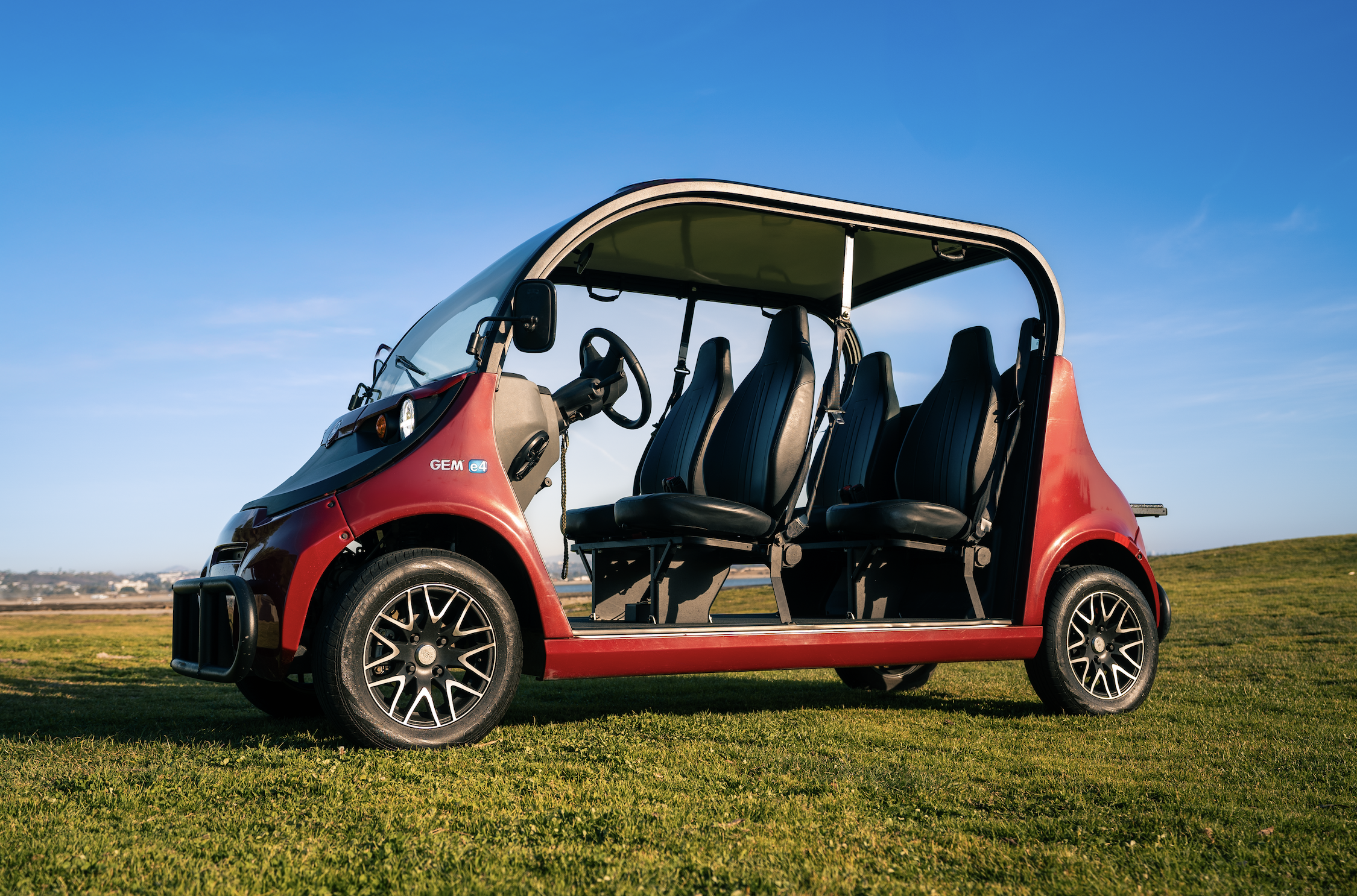 Red electric golf cart with four black seats parked on a grassy field under a clear blue sky.