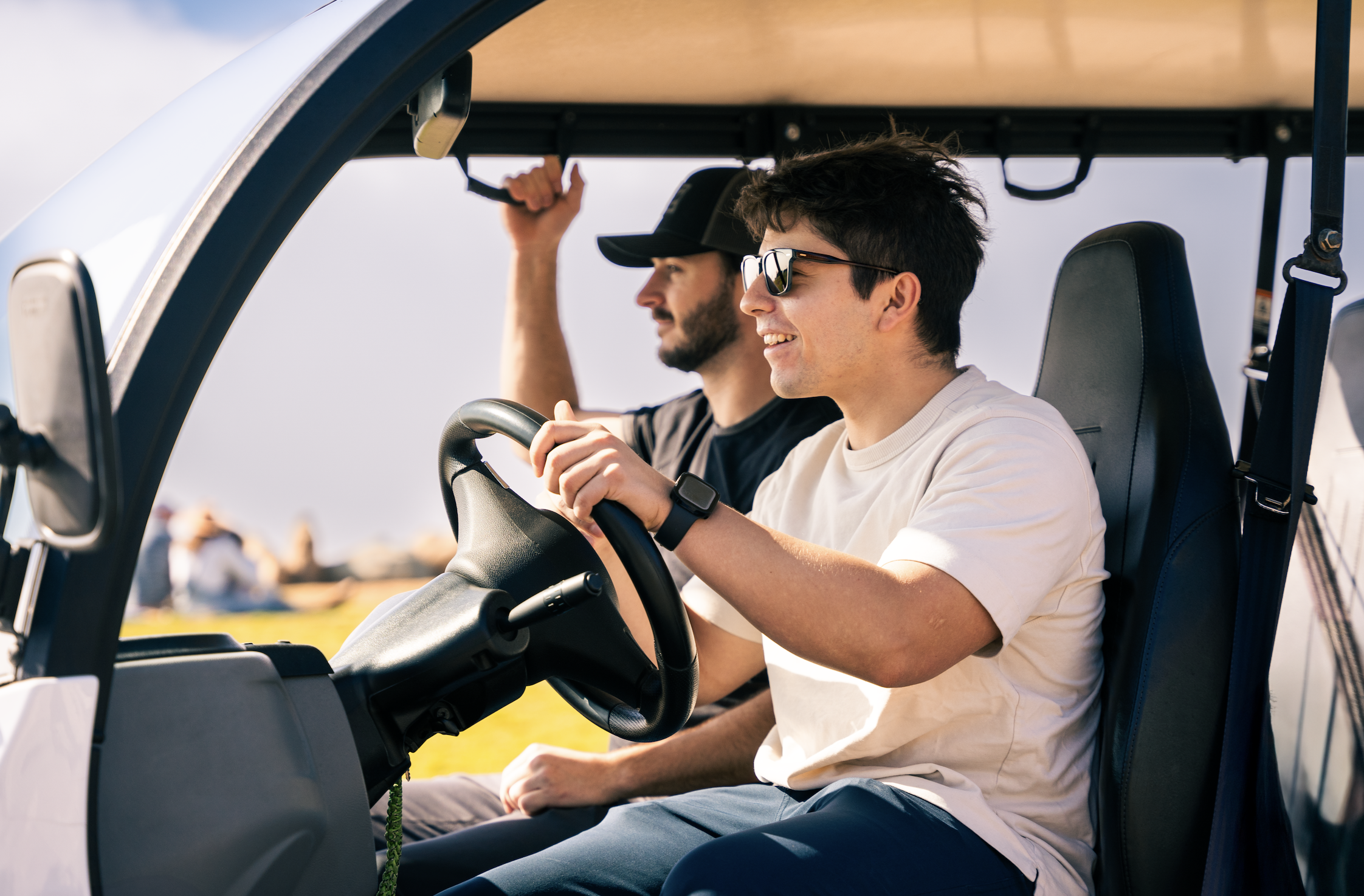Two young men smiling, one wearing sunglasses, driving a golf cart outdoors on sunny day.