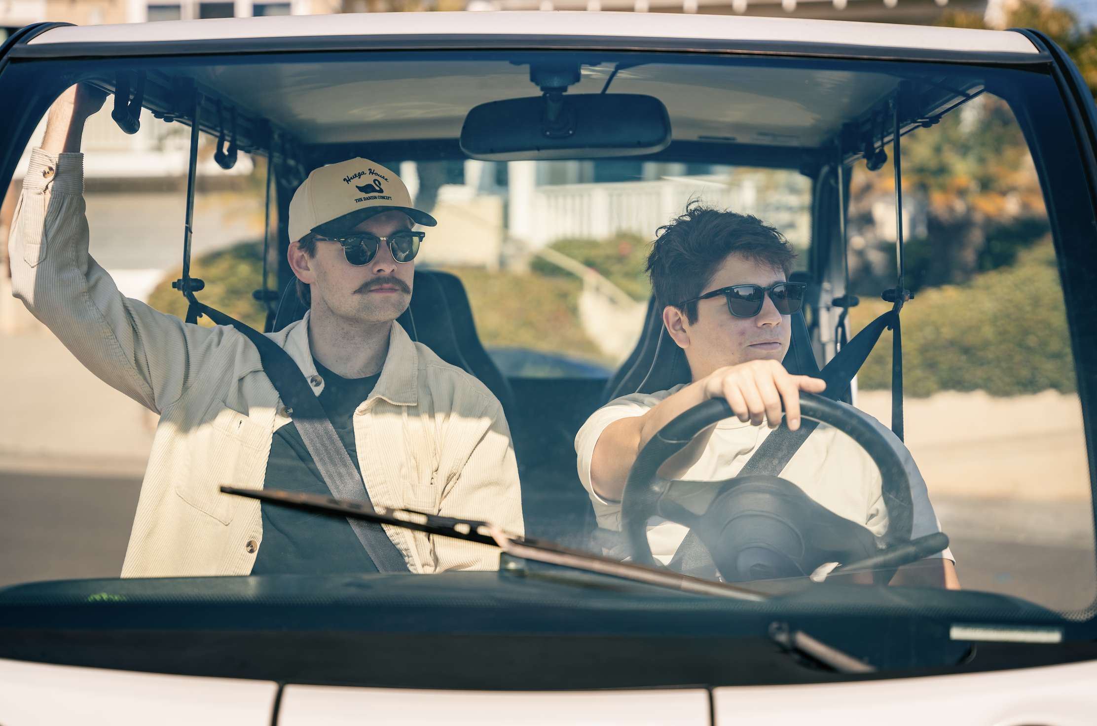 Two men with sunglasses riding and driving a golf cart on a sunny day.