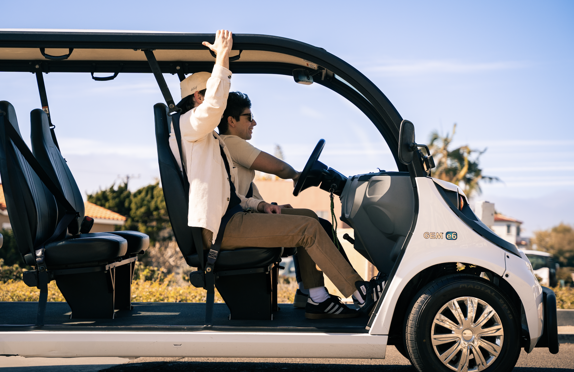 Two people riding in a small electric vehicle with open sides on a sunny day. One person is driving and the other is raising their hand, wearing sunglasses and casual clothing. Houses and trees are visible in the background.