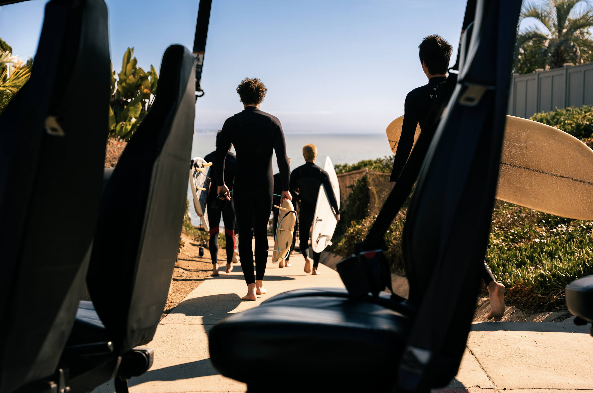 Surfers walking towards the ocean with surfboards, seen from inside a vehicle with seats in the foreground.