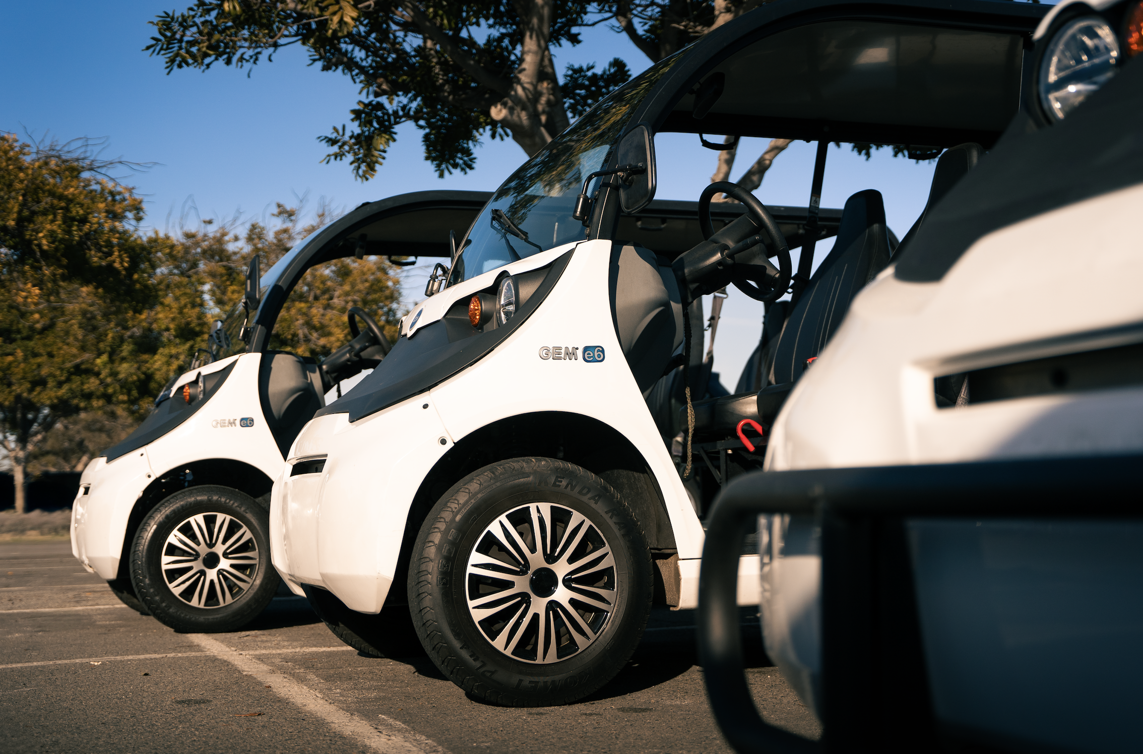 Two white GEM e6 electric vehicles parked side by side in a parking lot, with trees and a blue sky in the background.