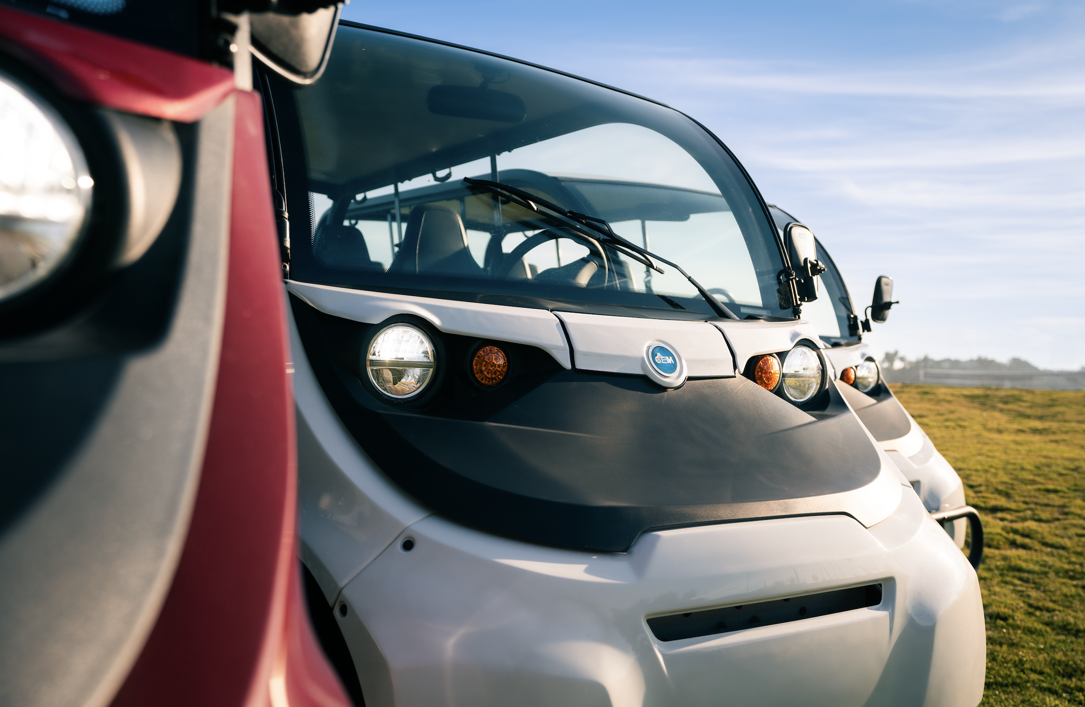 Close-up of two autonomous electric vehicles parked on grass under a clear blue sky, with the front of one vehicle visible on the left and the frontal view of the other vehicle in the center.