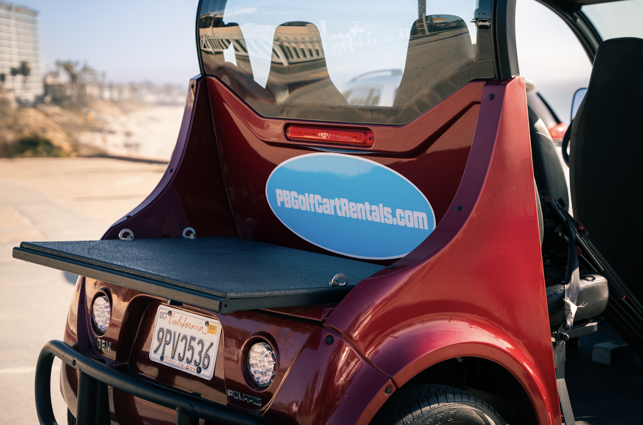 A red golf cart with a blue oval sticker that says 'PBGolfCartRentals.com' on the back, parked outdoors with a blurred background of buildings and trees.