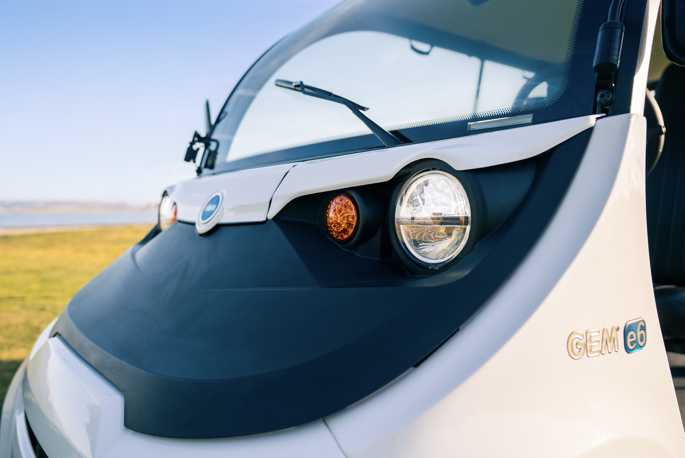 Close-up of a white GEM e6 electric vehicle, showing its front with headlight, turn signal, windshield, and the GEM e6 badge on the side.