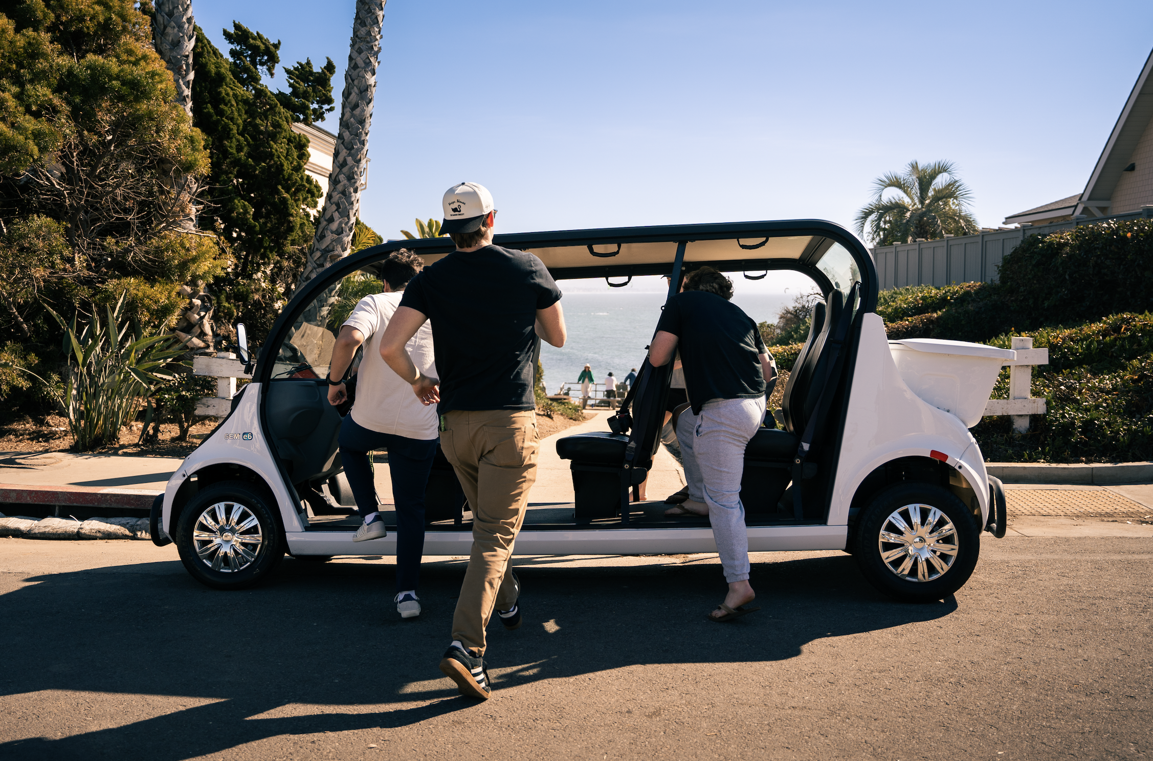 Group of people around a white golf cart parked on a scenic coastal road with trees, shrubbery, and ocean in the background during daytime.