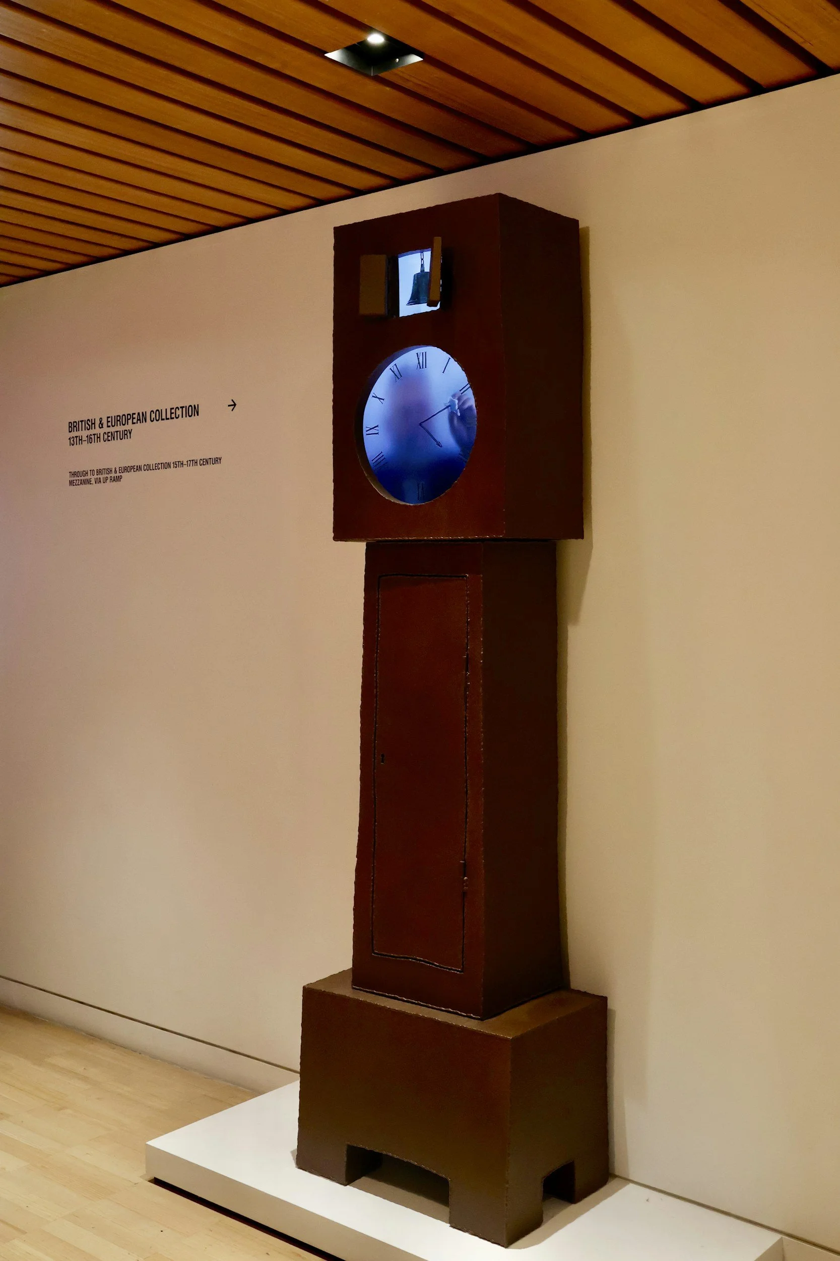 A tall, wooden grandfather clock with a glass face showing Roman numerals and clock hands, standing on a base in a museum or gallery setting.