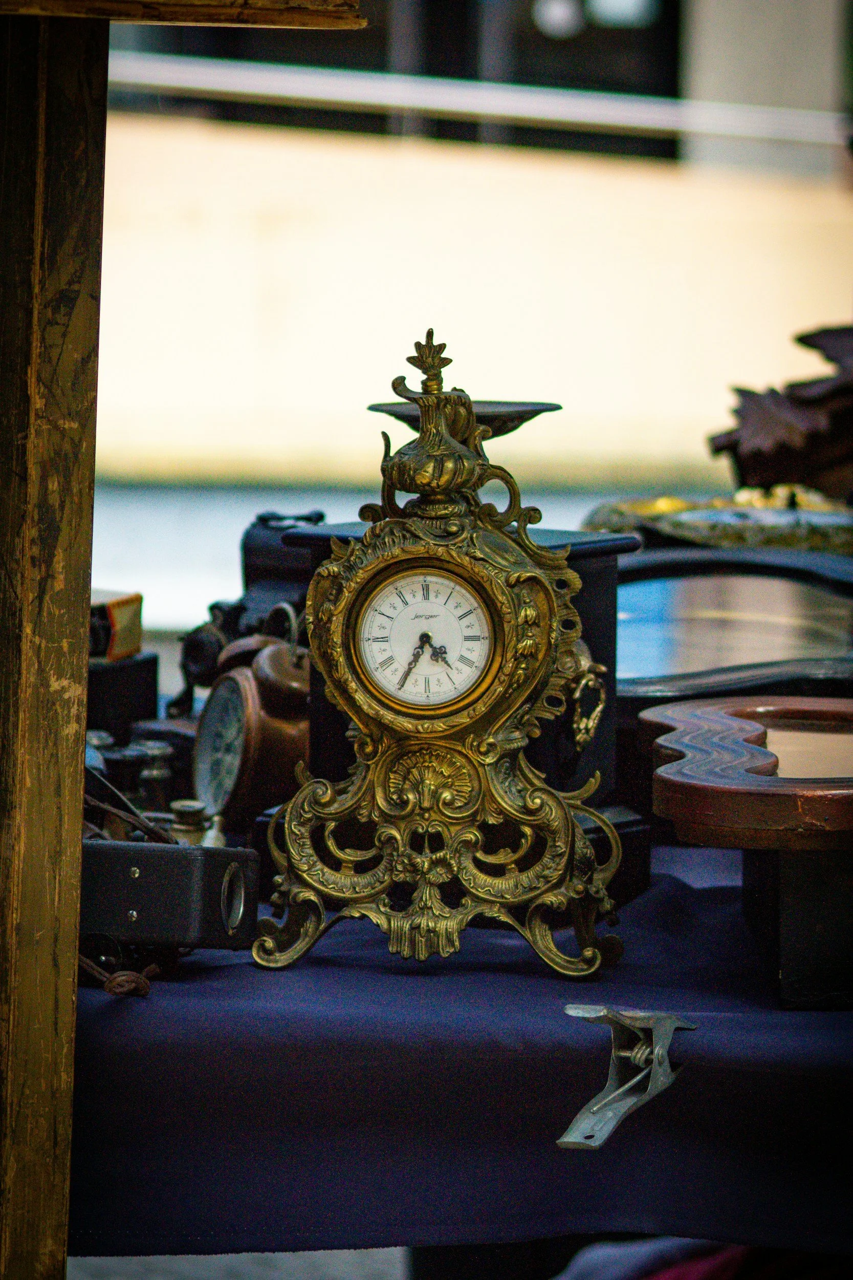 An ornate antique clock sits on a table at an outdoor market, surrounded by various other vintage items.