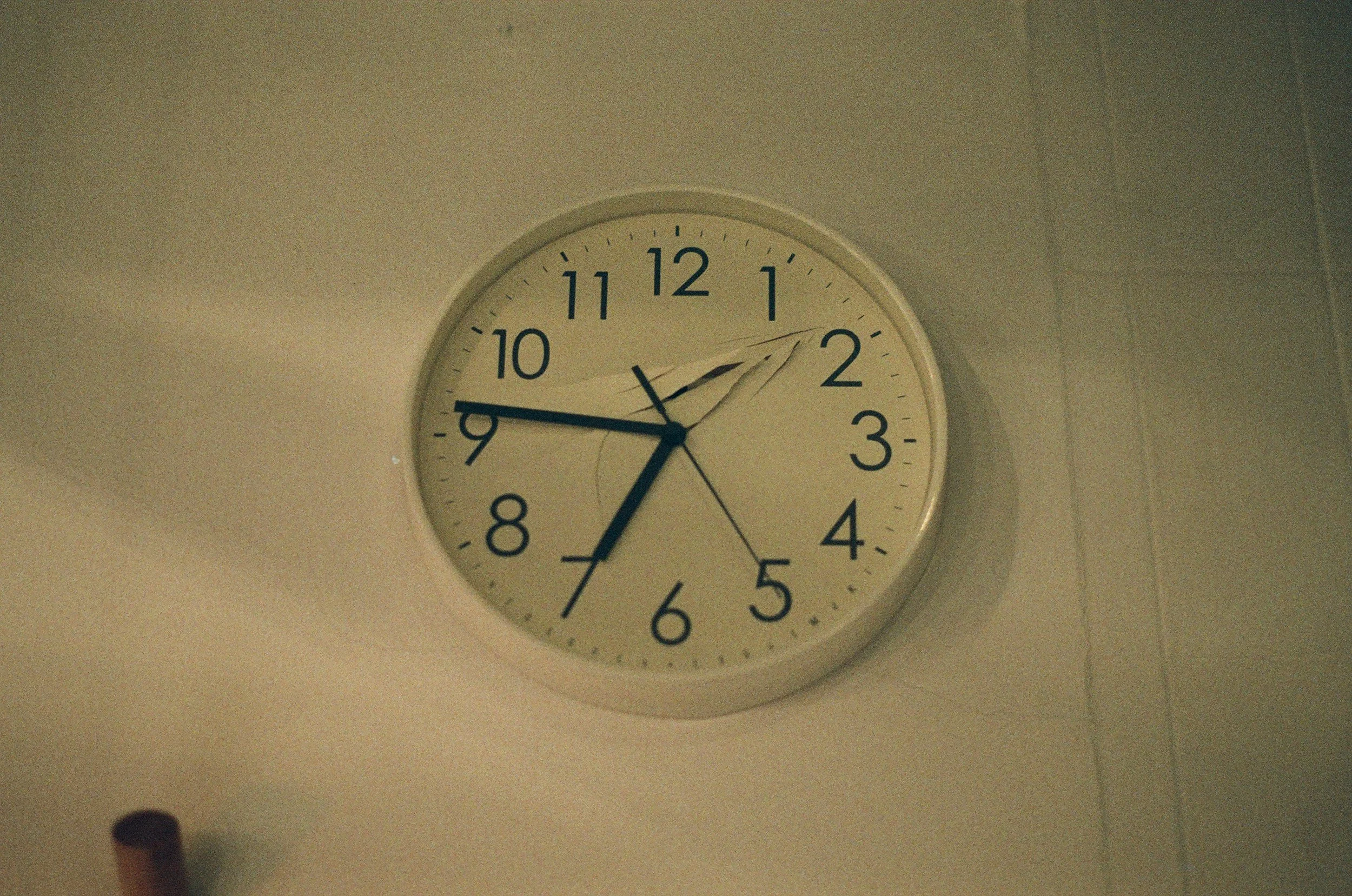 An analog wall clock showing the time as 1:43 with some cracks on the glass and mounted on a plain wall.