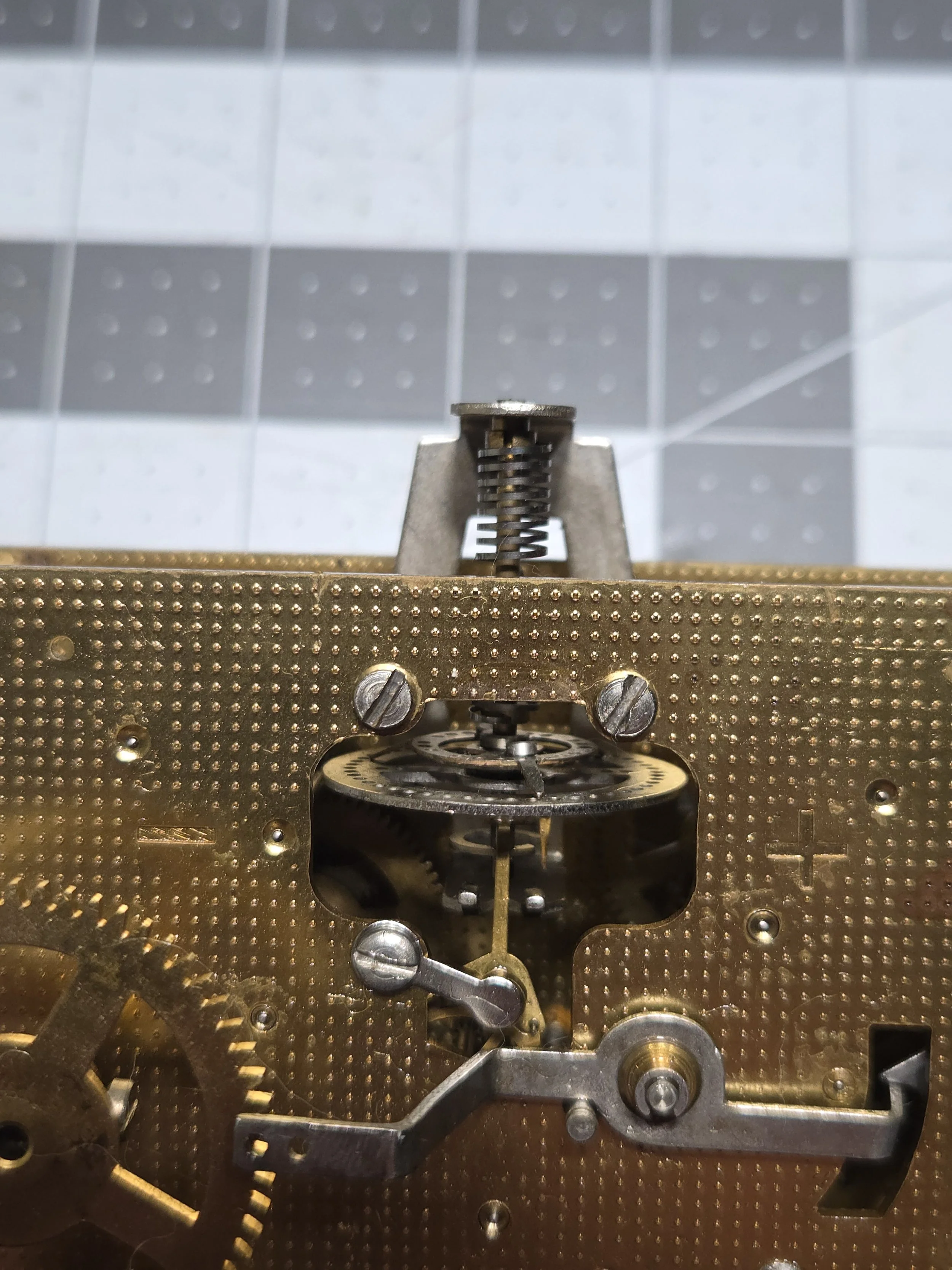 Close-up of the inner mechanical workings of a vintage clock or watch, featuring gears, screws, and a metal plate with a textured surface.