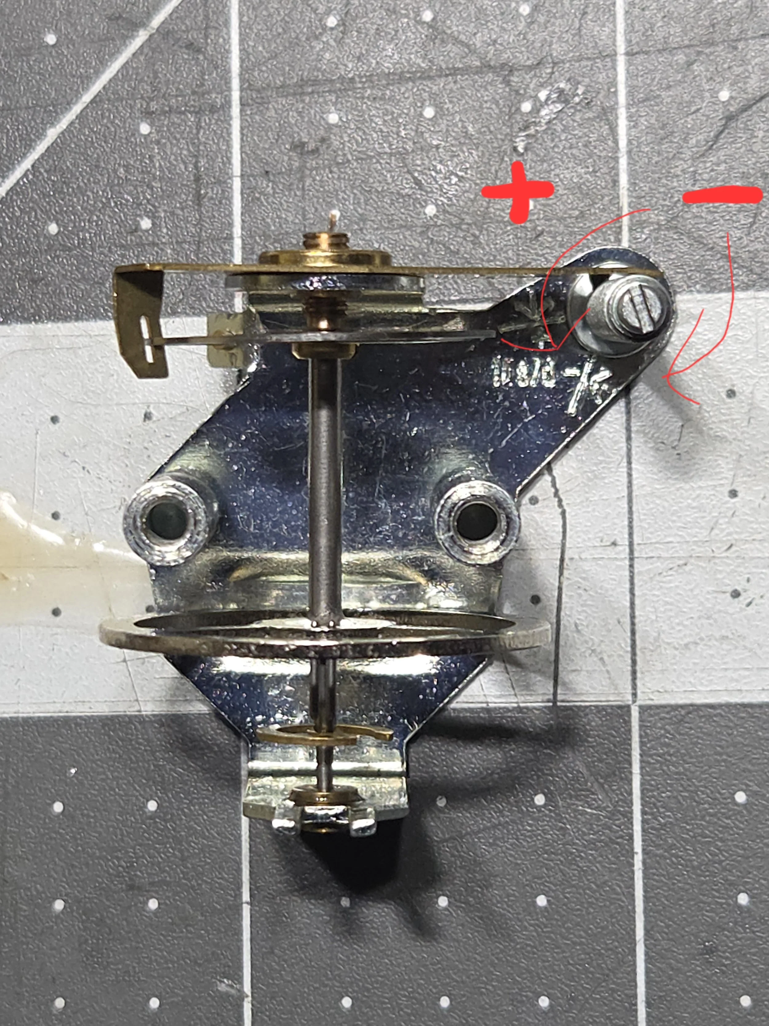 Metal mechanical part with screws and a rod, mounted on a pegboard with markings and annotations in red pen.