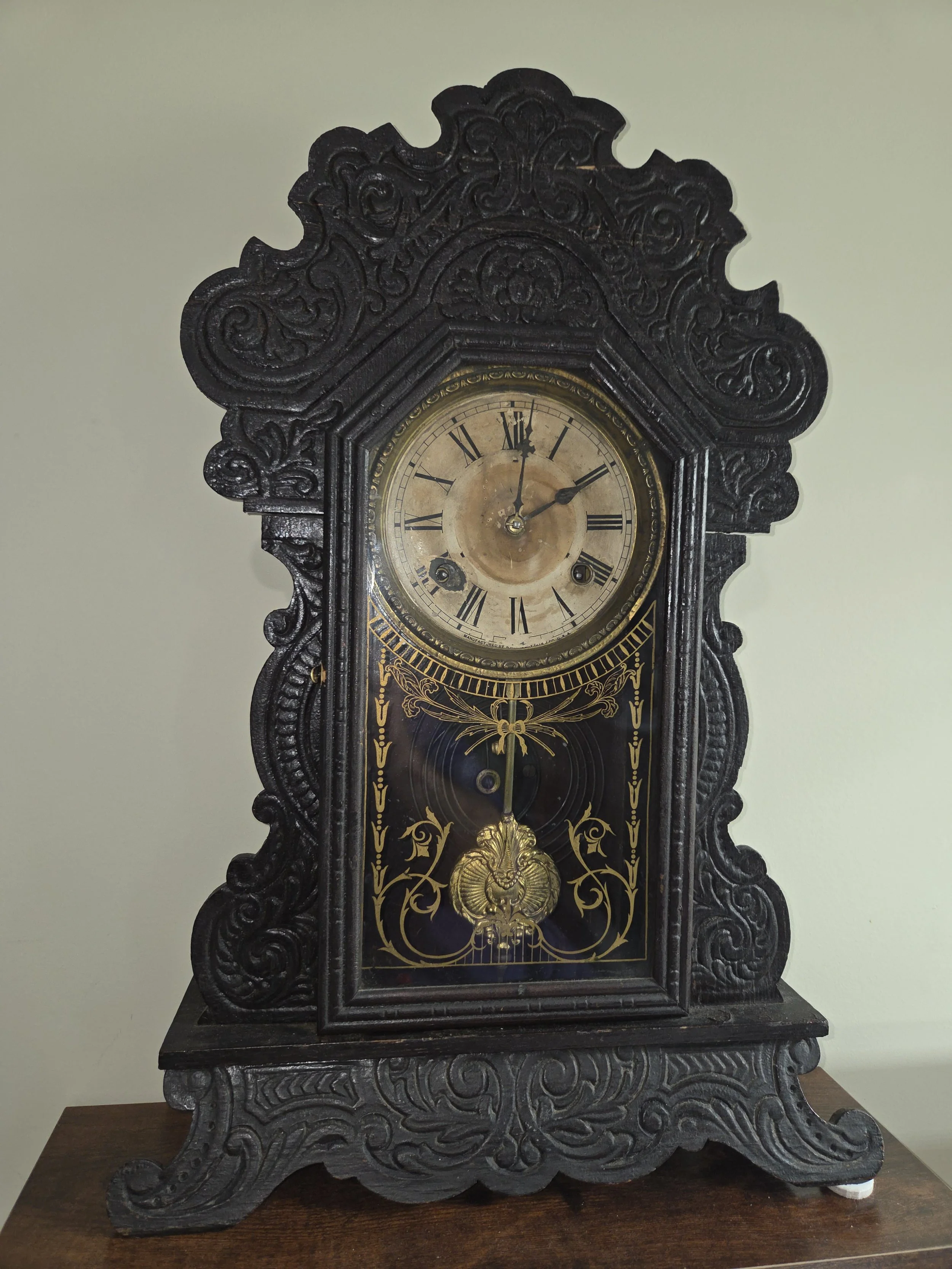 An antique black wooden clock with ornate carvings, Roman numerals on a gold clock face, and decorative gold detailing on the glass panel.