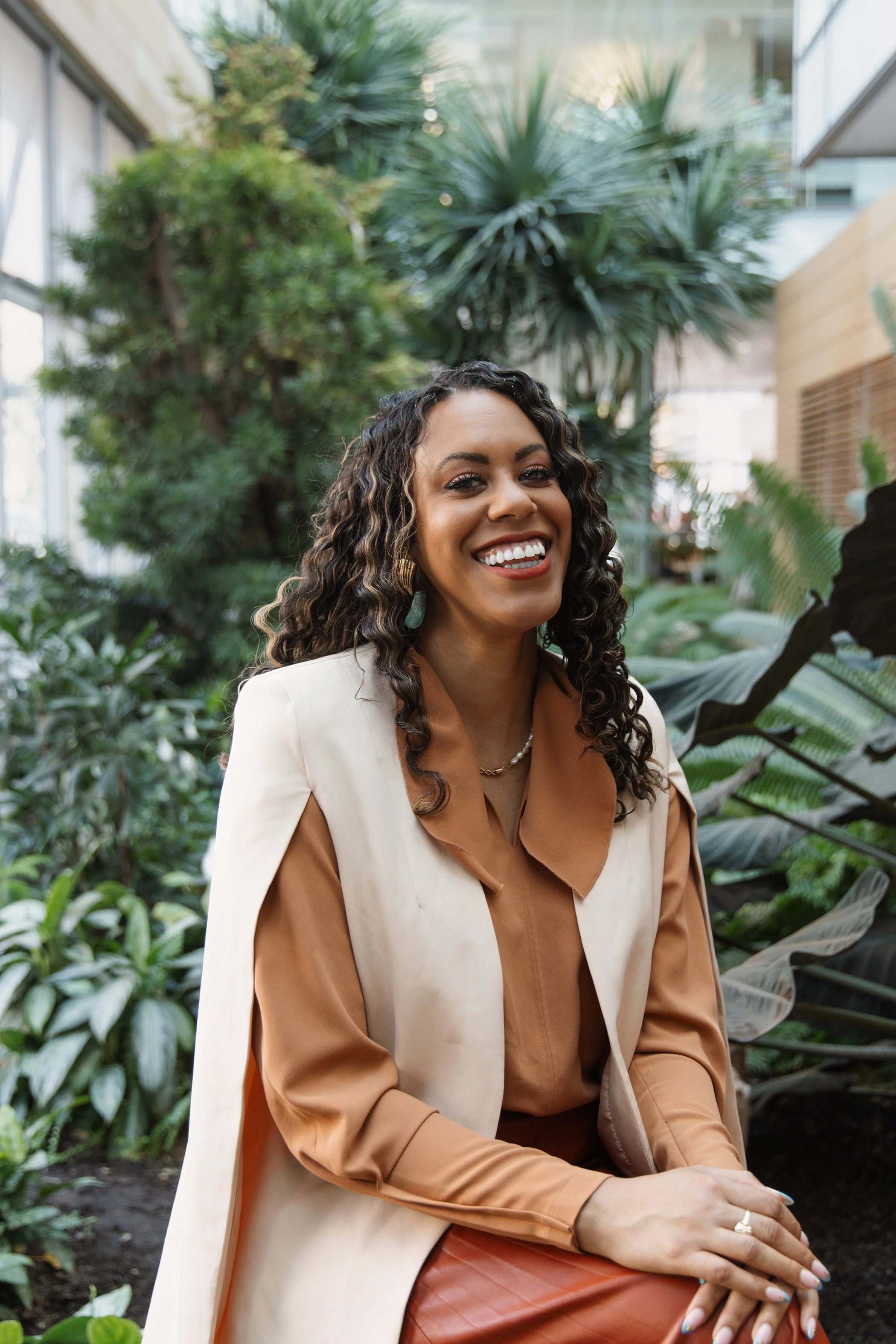 A smiling woman seated in a lush indoor garden with various green plants and trees.