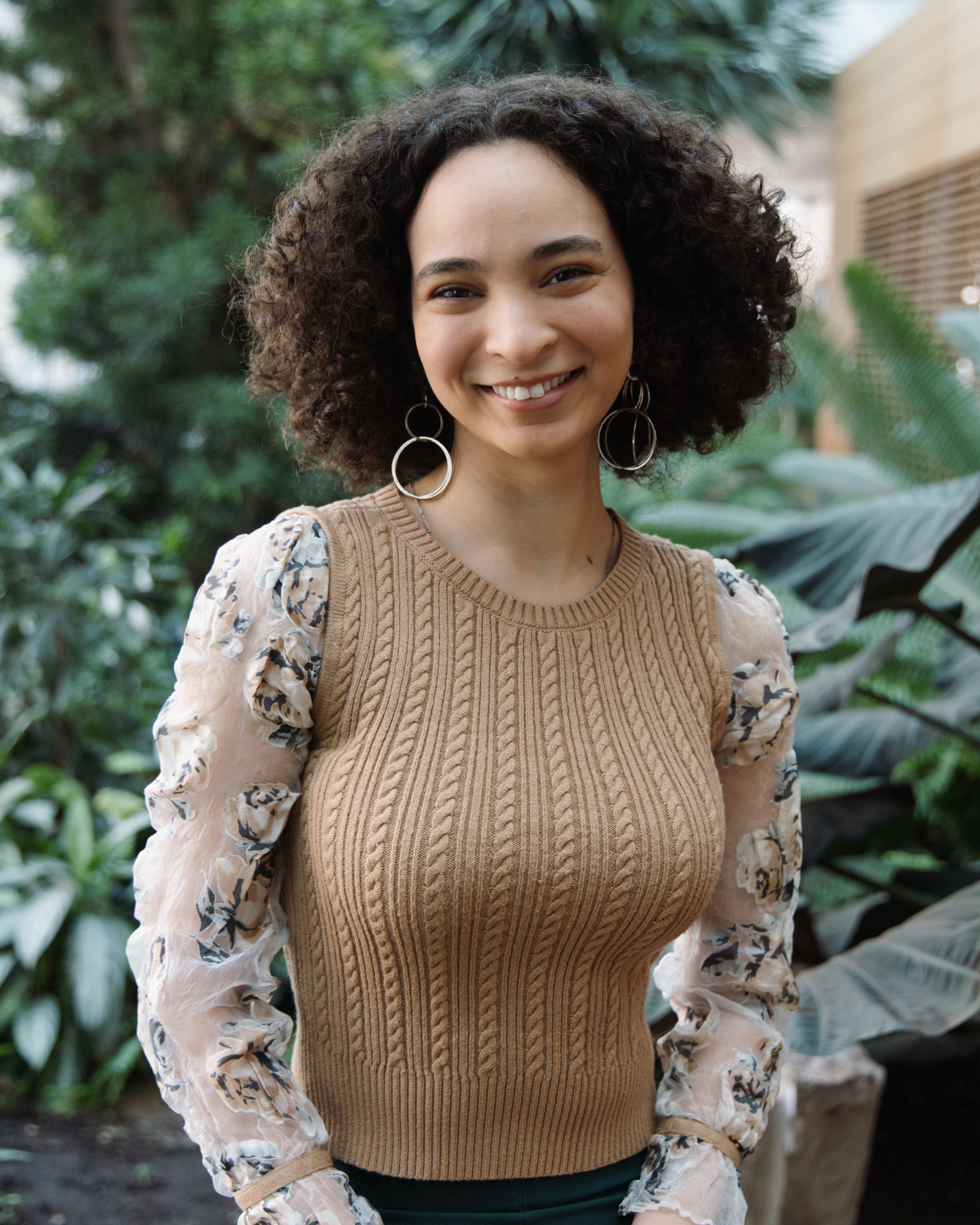 A young woman with curly hair, wearing a brown knit sweater with floral sheer sleeves, smiling in a lush indoor garden.