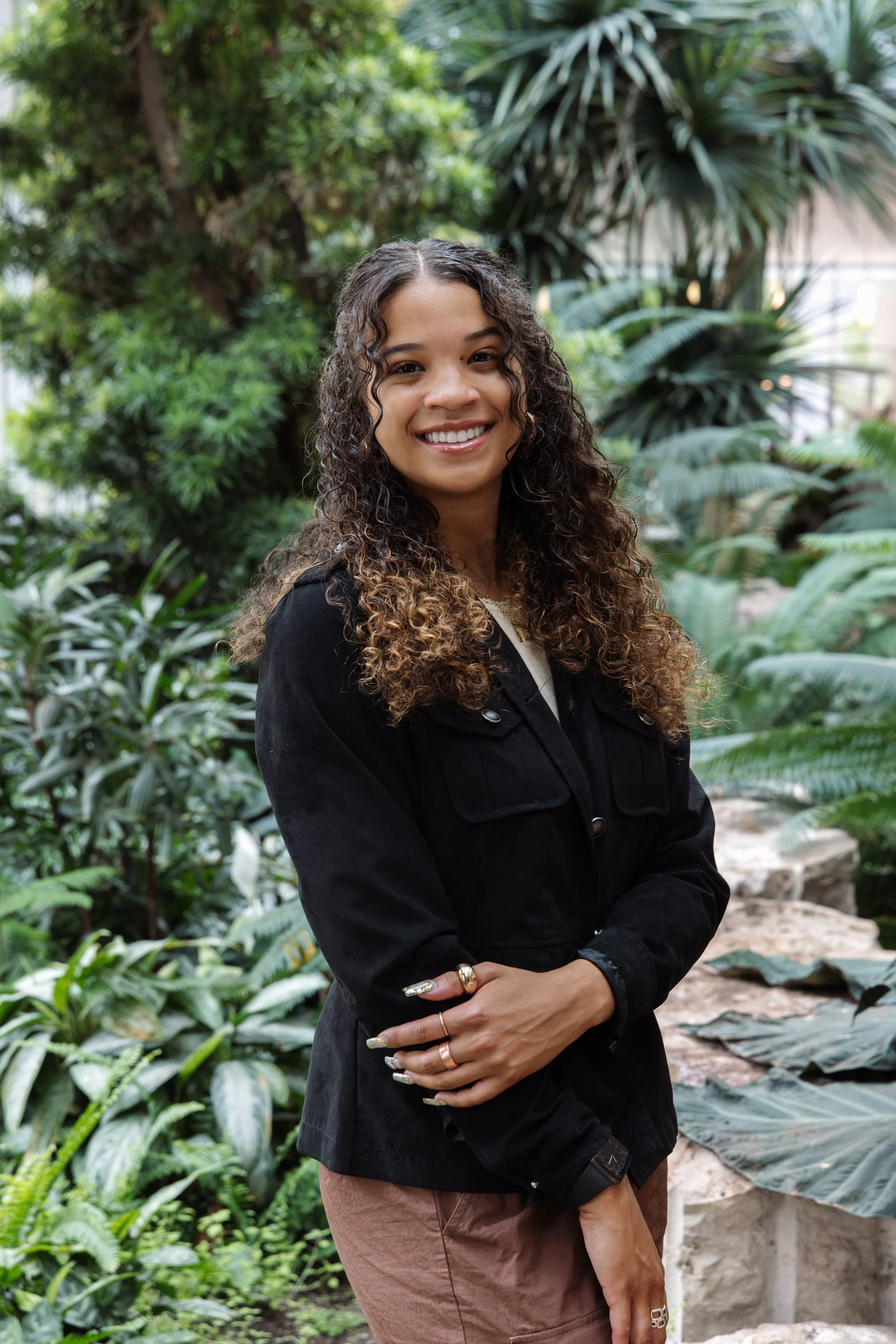 A woman with curly brown hair smiling in a lush indoor garden with various green plants.