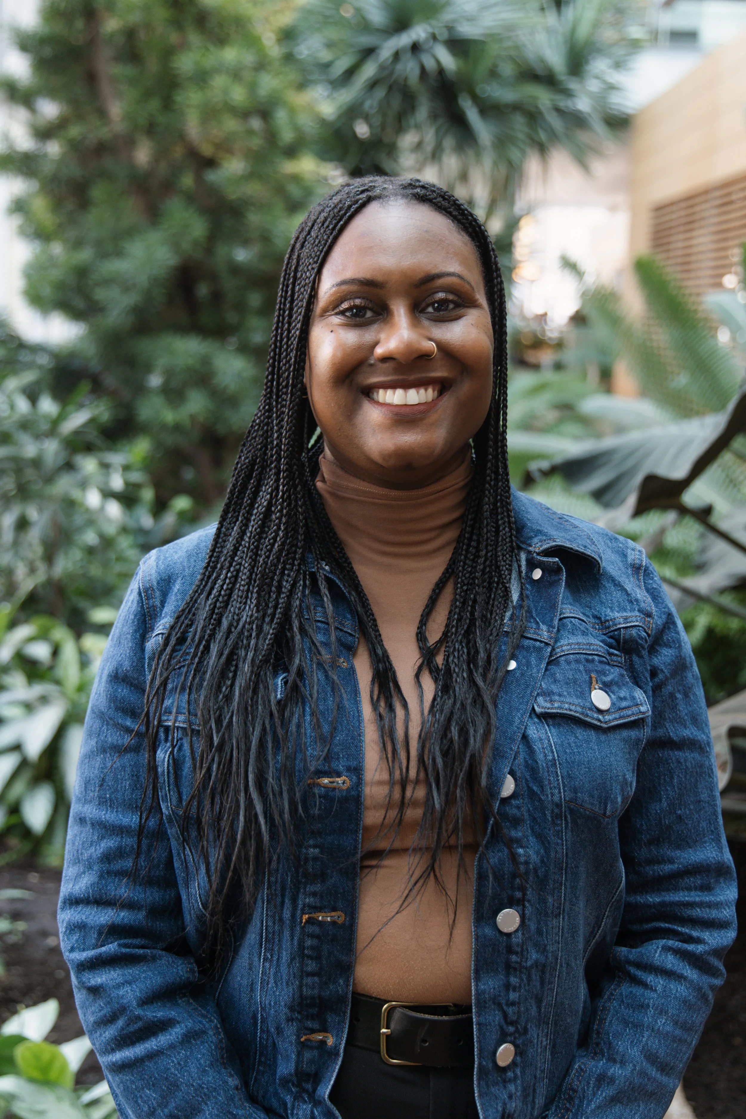 A woman with long braided hair, wearing a denim jacket over a brown turtleneck, smiling in a lush, green outdoor setting.