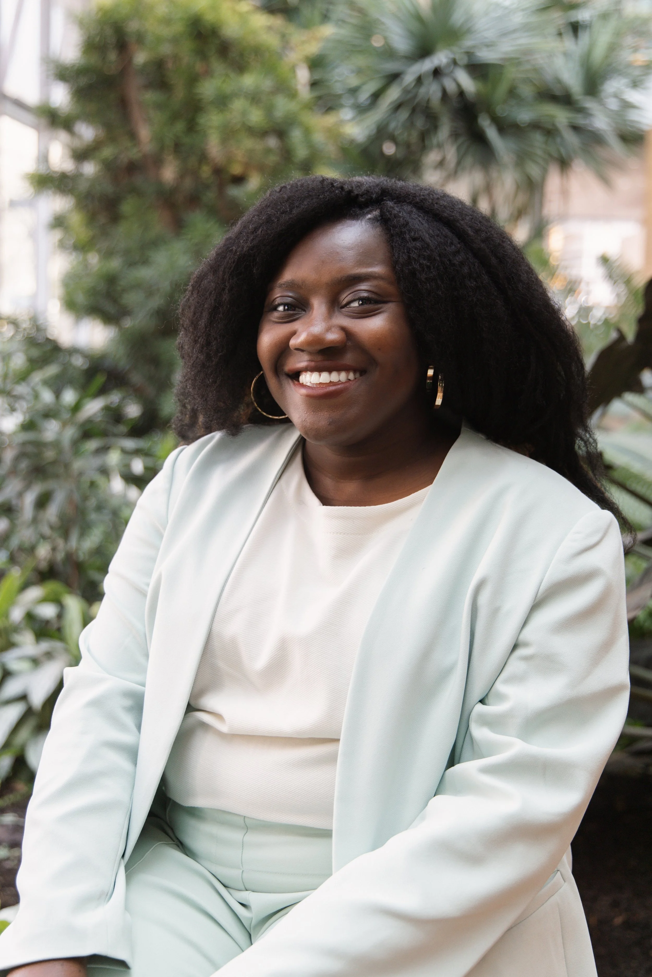A woman with dark skin, black curly hair, and wearing a light mint green blazer and matching pants, smiling while sitting outdoors with green plants in the background.