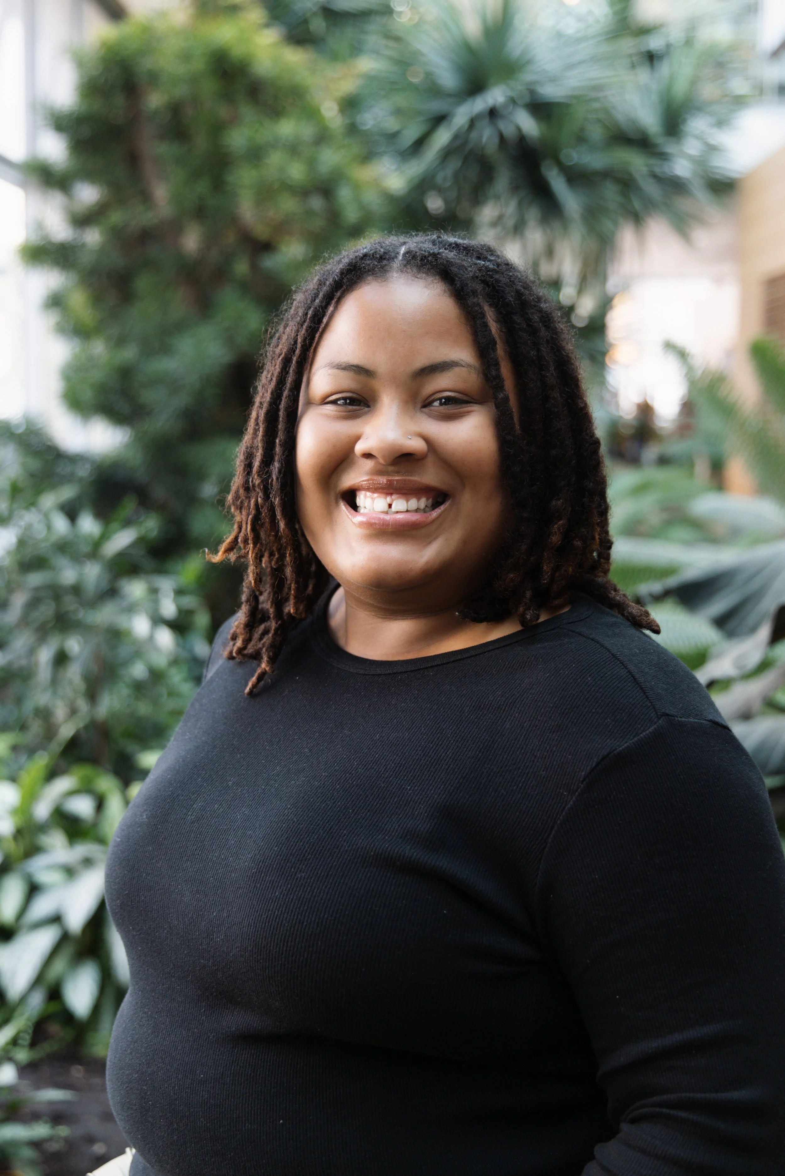 A smiling woman with dreadlocks wearing a black sweater standing in a lush green indoor garden.
