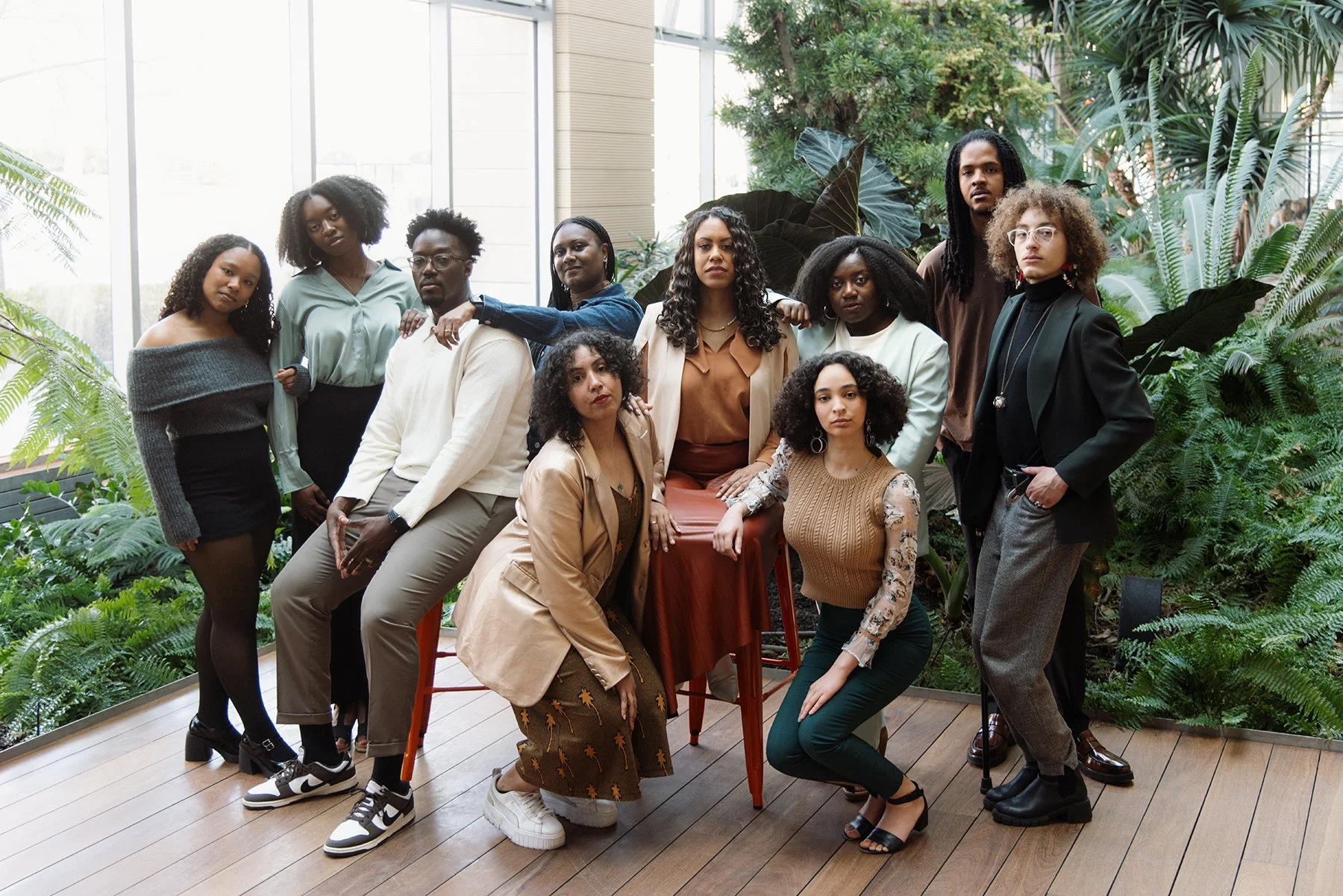 A diverse group of eleven people, eleven women and one man, posing together in an indoor botanical setting with lush green plants and wooden floors.
