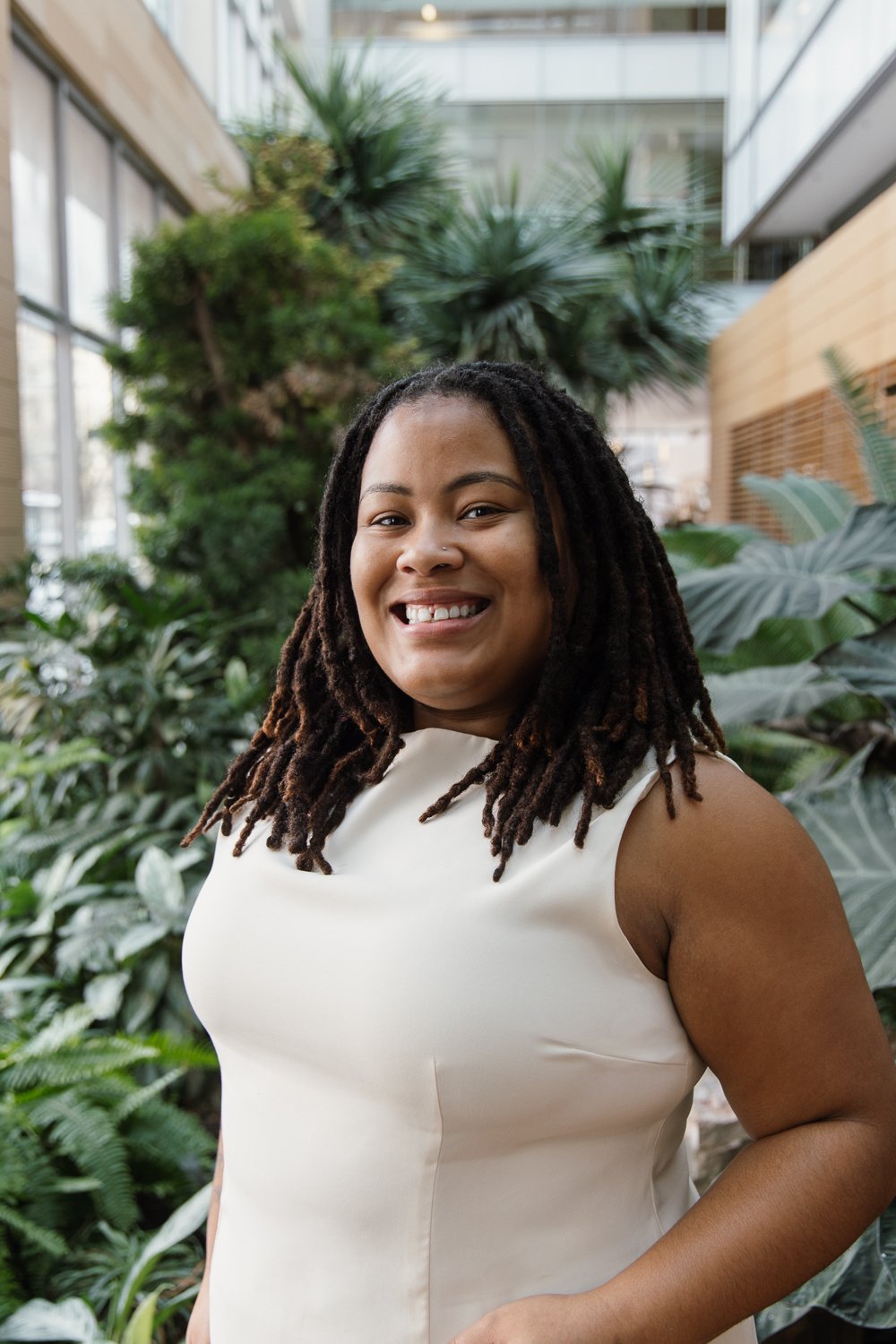 A smiling woman with dreadlocks wearing a black sweater standing in a lush green indoor garden.