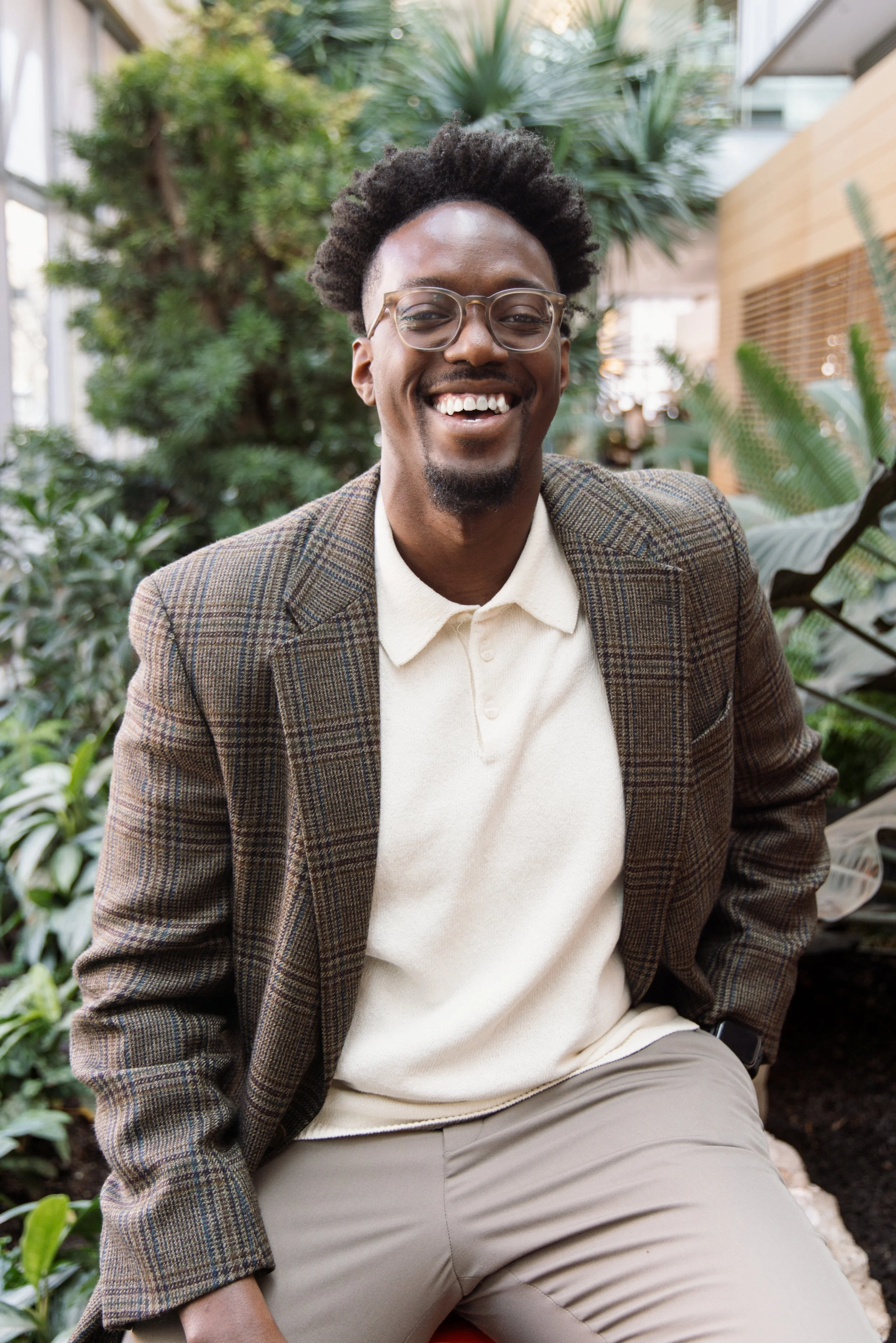 A smiling man with glasses and a beard, wearing a plaid blazer over a cream polo shirt, sitting in a lush indoor garden.