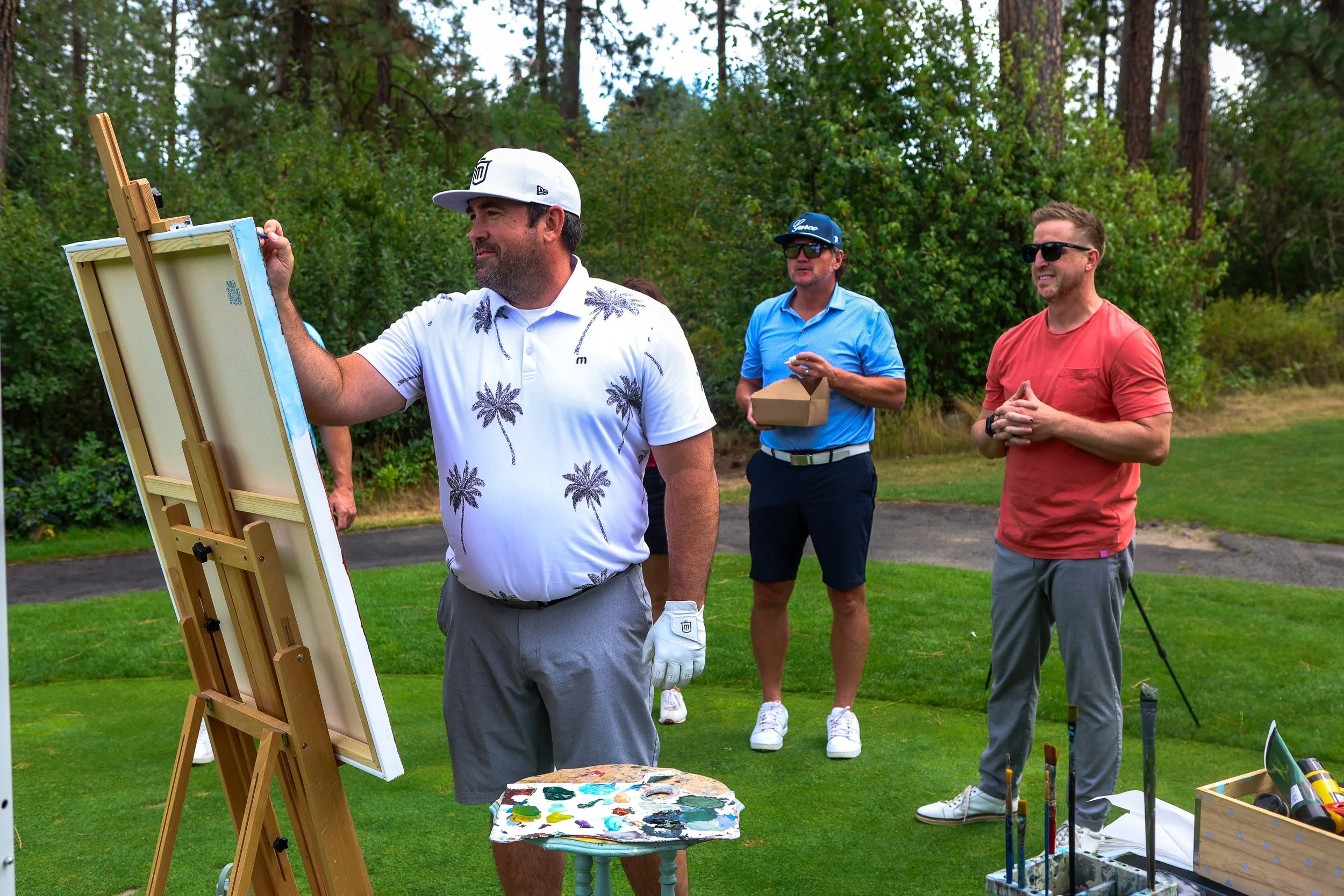 Man in a white shirt and gray shorts signing a painting while two other people watch.