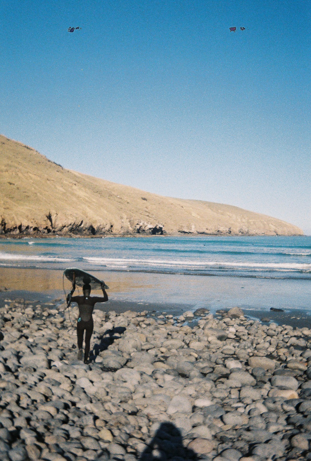 A person walking on a rocky beach holding a surfboard above their head, with ocean waves and a hillside in the background.