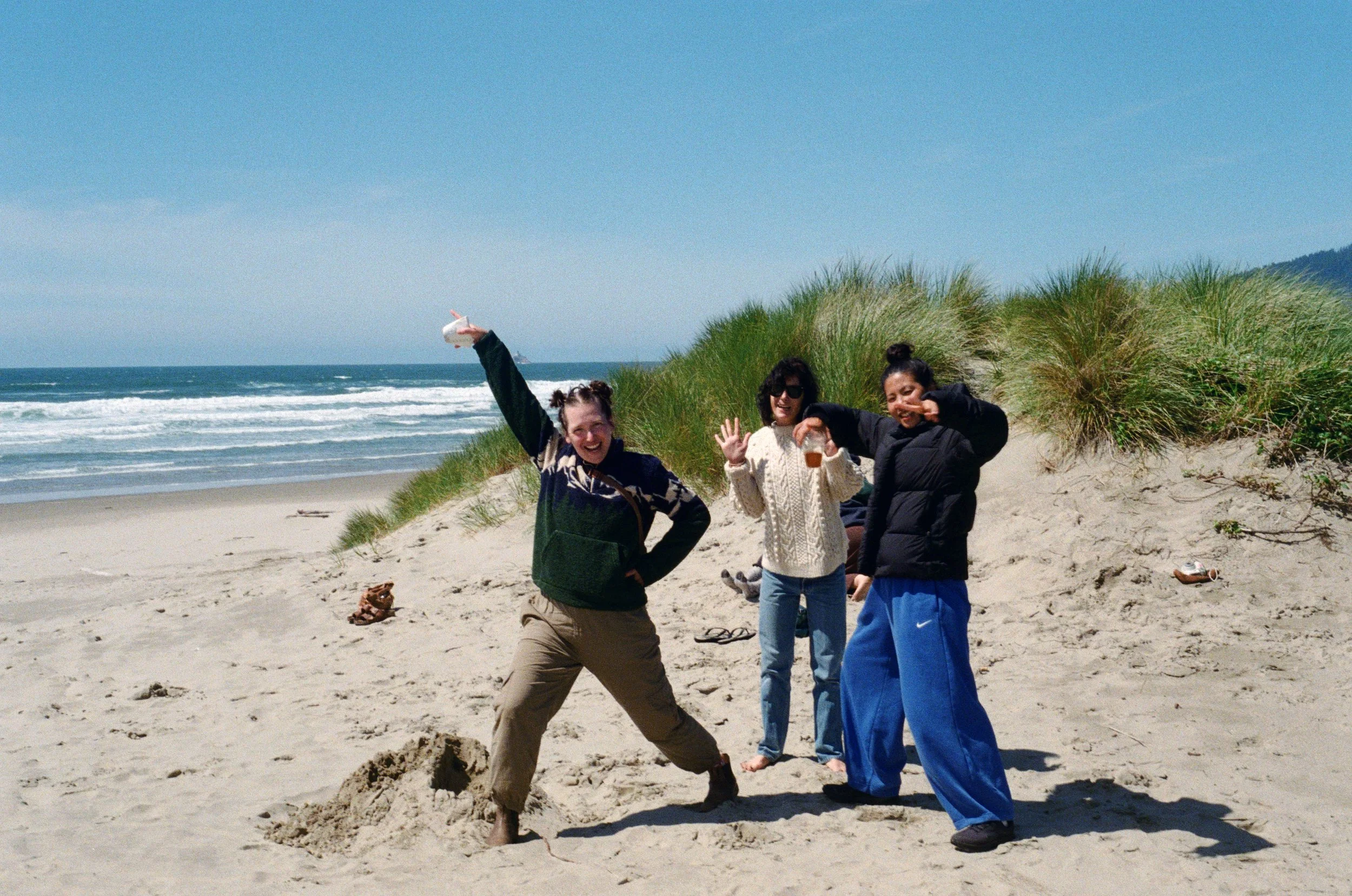Three women at the beach, one of whom is making a snow angel in the sand while the other two pose and smile.