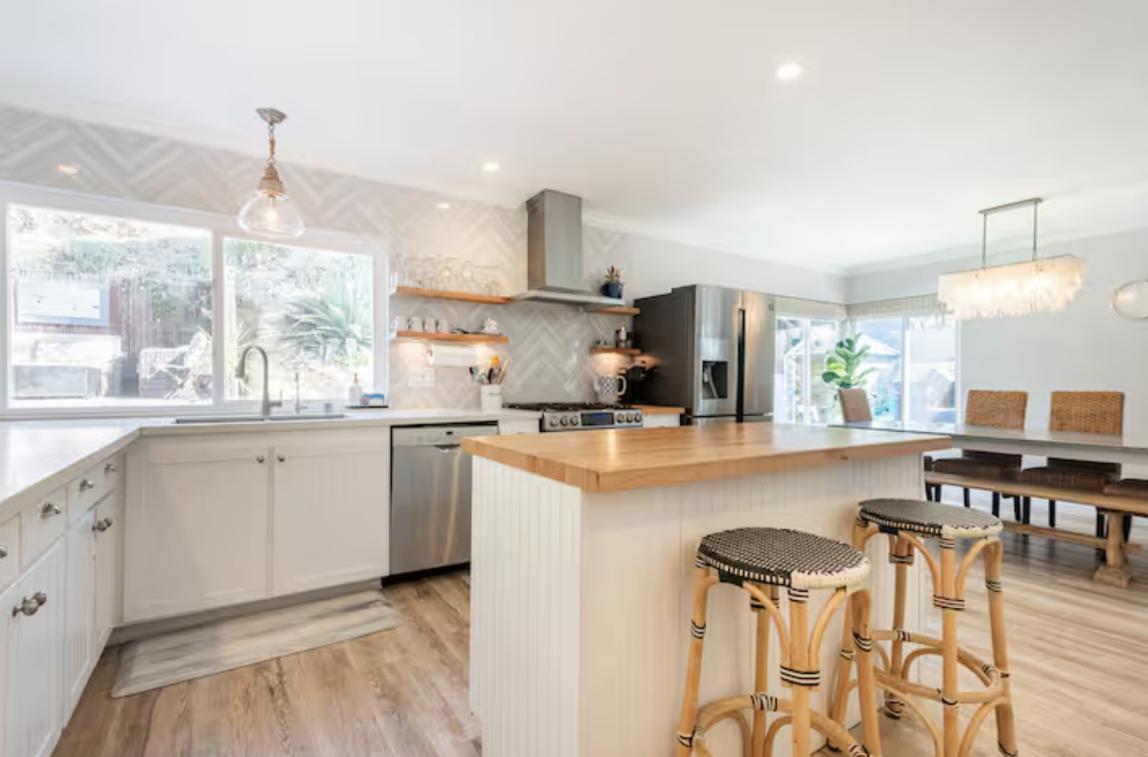 Bright kitchen with white cabinets, wooden accents, stainless steel appliances, and a large island with barstools, adjacent to a dining area with chairs, natural light, and indoor plants.