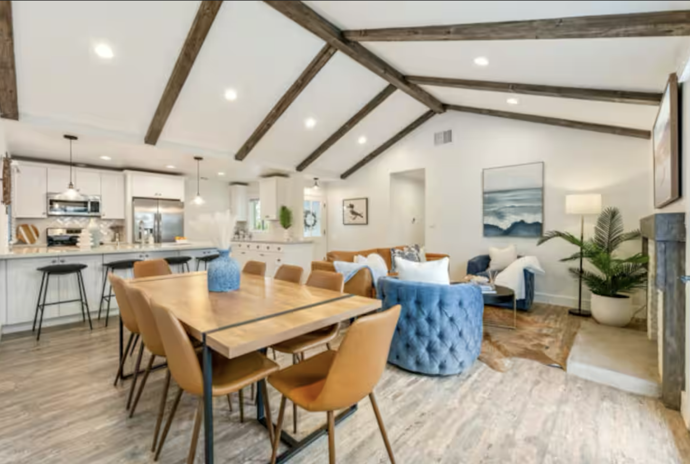 Open-concept living space with a kitchen in the background, featuring white cabinetry, a large island, and black barstools. The living area has a wooden dining table, tan chairs, blue and white cushioned armchairs, potted plants, and textured wood ceiling beams.