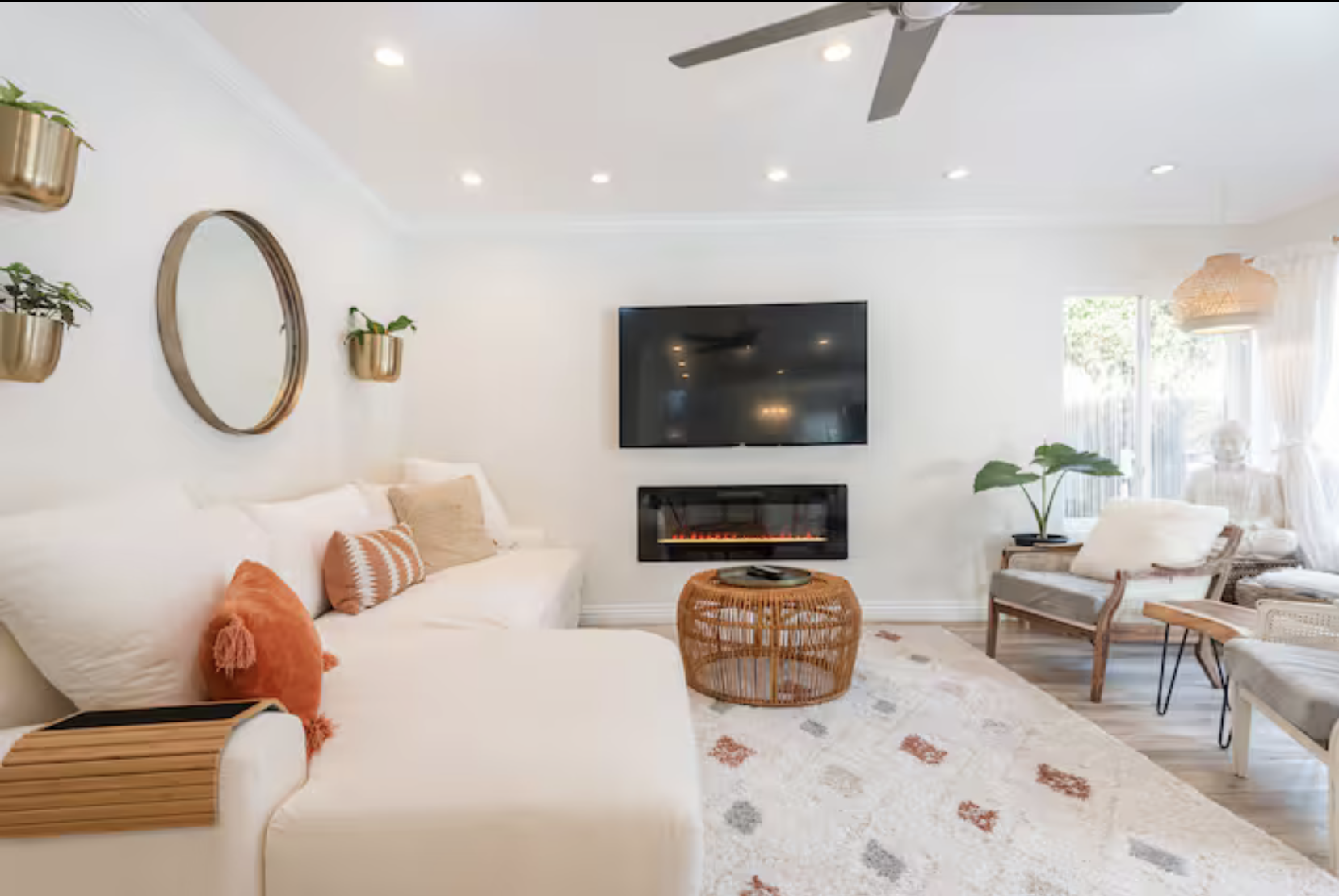Bright living room with white walls and a ceiling fan, featuring a wall-mounted TV above an electric fireplace. There is a cream sofa decorated with orange and beige pillows, a wicker coffee table, and several armchairs near large windows with sheer curtains. Wall planters with plants and a round mirror adorn the wall.