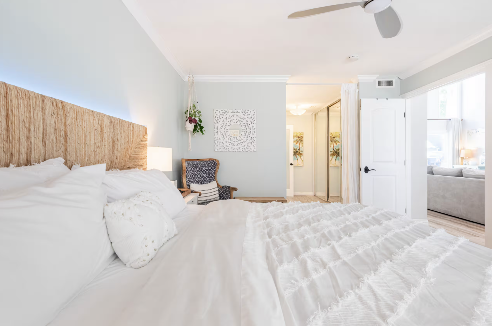 A bedroom with a large bed that has a textured beige headboard, white bedding, and pillows. There is a small nightstand with a lamp, a patterned armchair with a cushion, and a wall hanging with a plant. The room has a ceiling fan, a built-in closet with mirrored doors, and an open door leading to another bright living space with large windows, a grey sofa, and tropical-themed artwork.