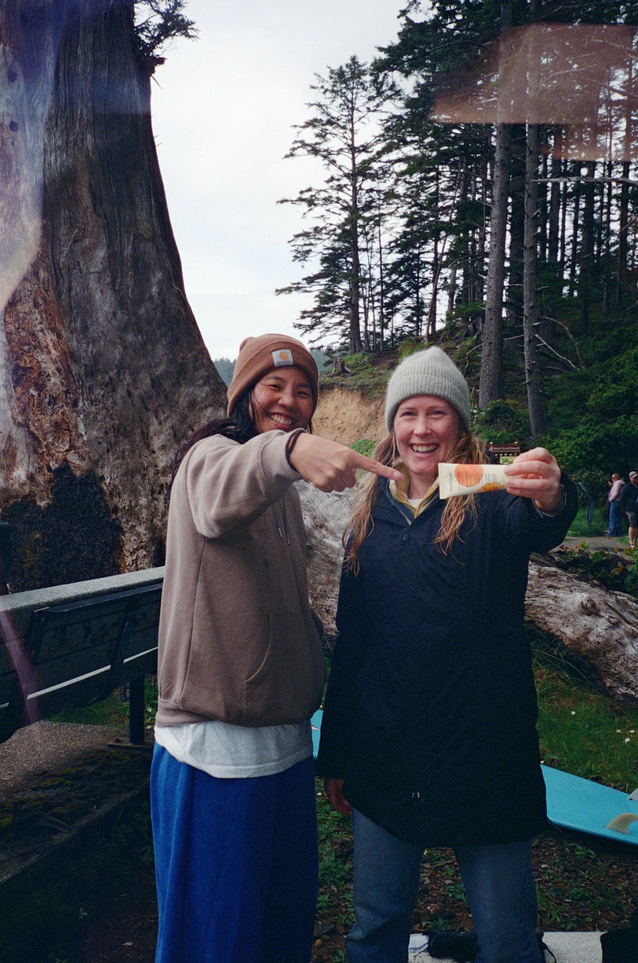 Two smiling women outdoors, one pointing at a tube of sunscreen the other holding it, near a large fallen tree and forest background.