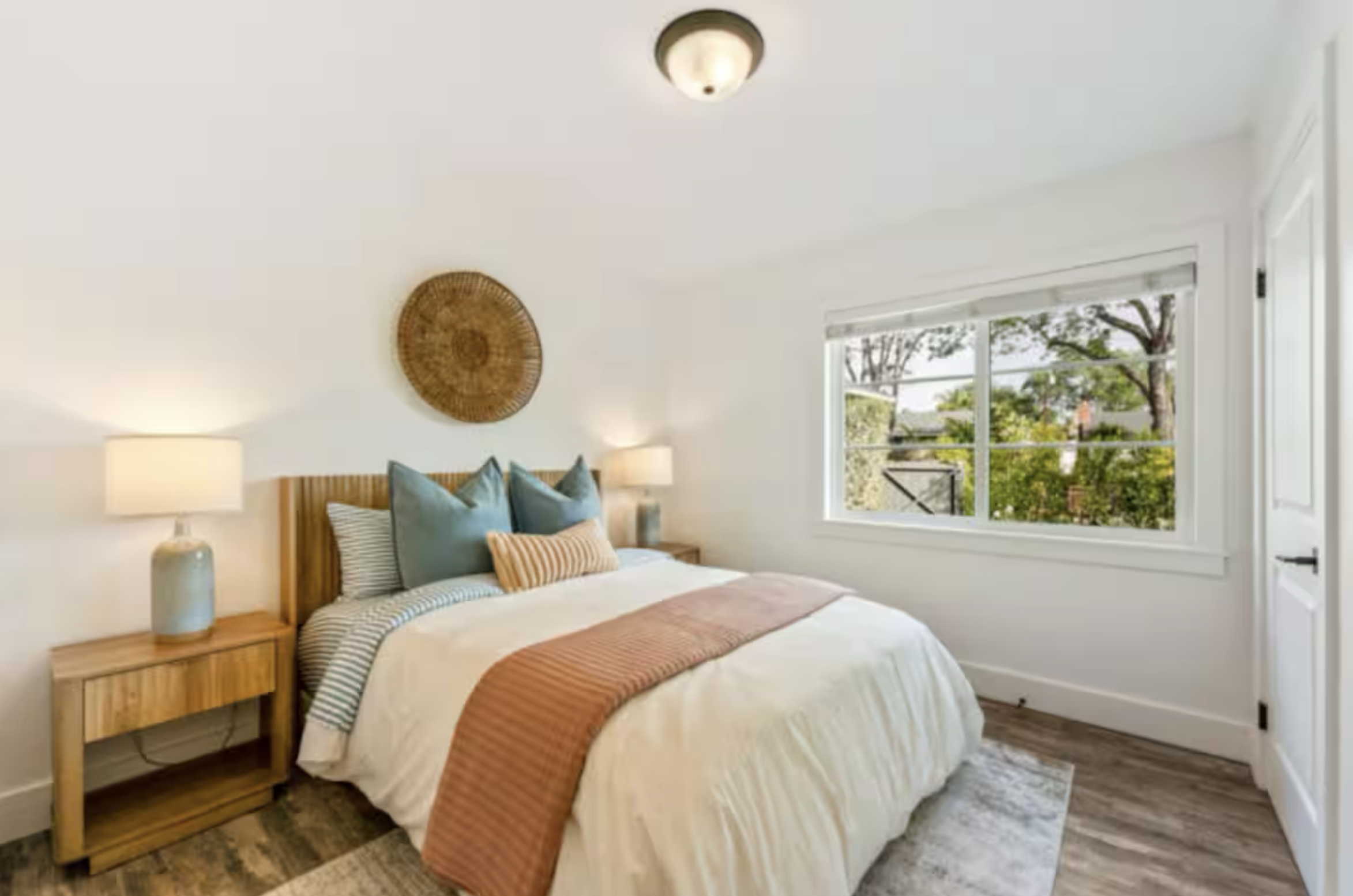 A bedroom with a wooden bed frame, beige bedding, and multiple blue and beige pillows. There are two wooden nightstands with matching lamps. A woven circular wall hanging decorates the wall above the bed. A window with blinds reveals trees outside.