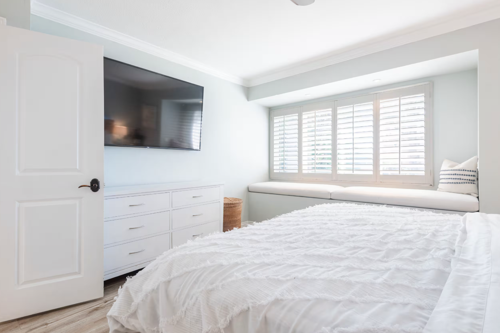 A bright bedroom with a large window, built-in window seat with pillows, white dresser, and a wall-mounted flat-screen TV, with a bed in the foreground.