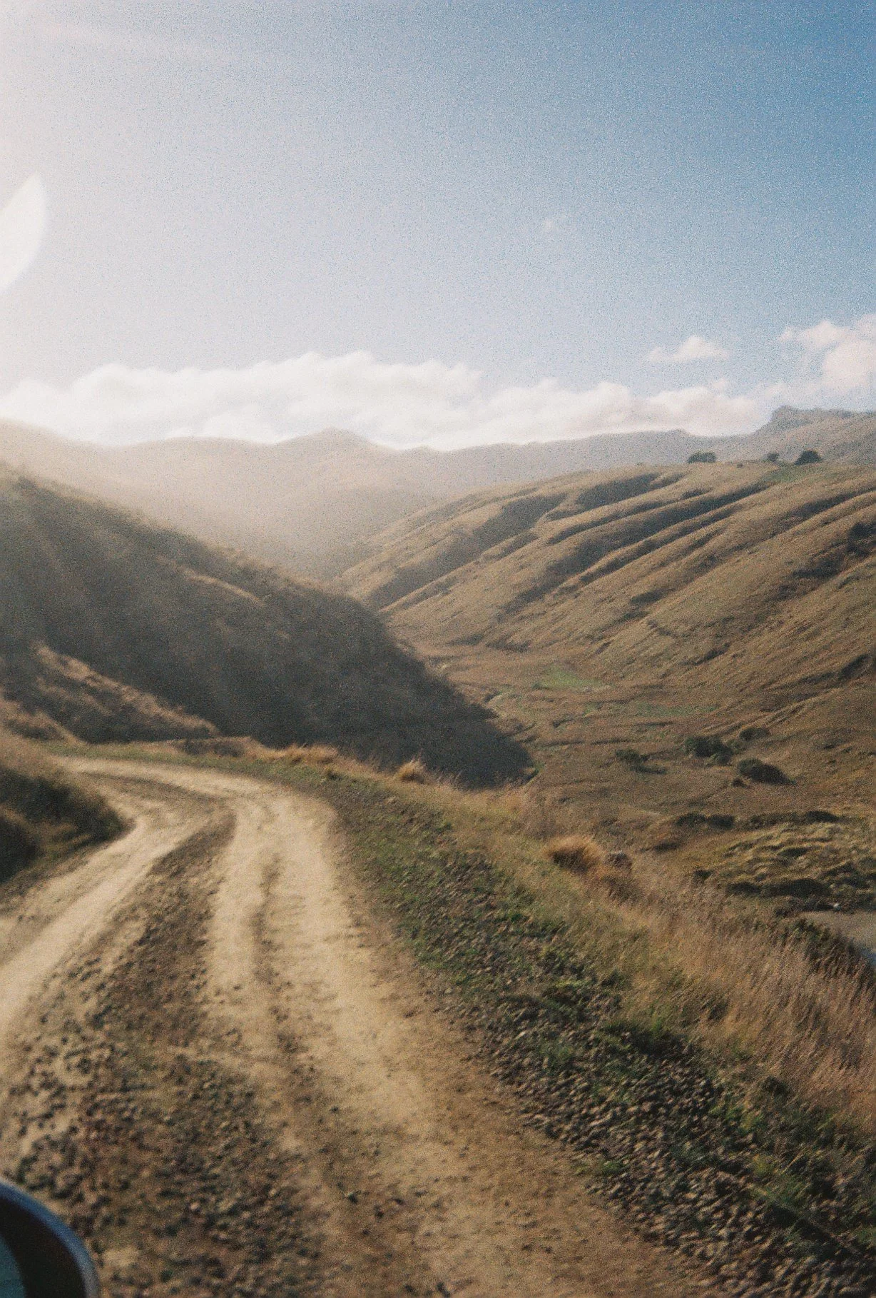 Dirt road winding through rolling hills with sparse vegetation under a cloudy sky.