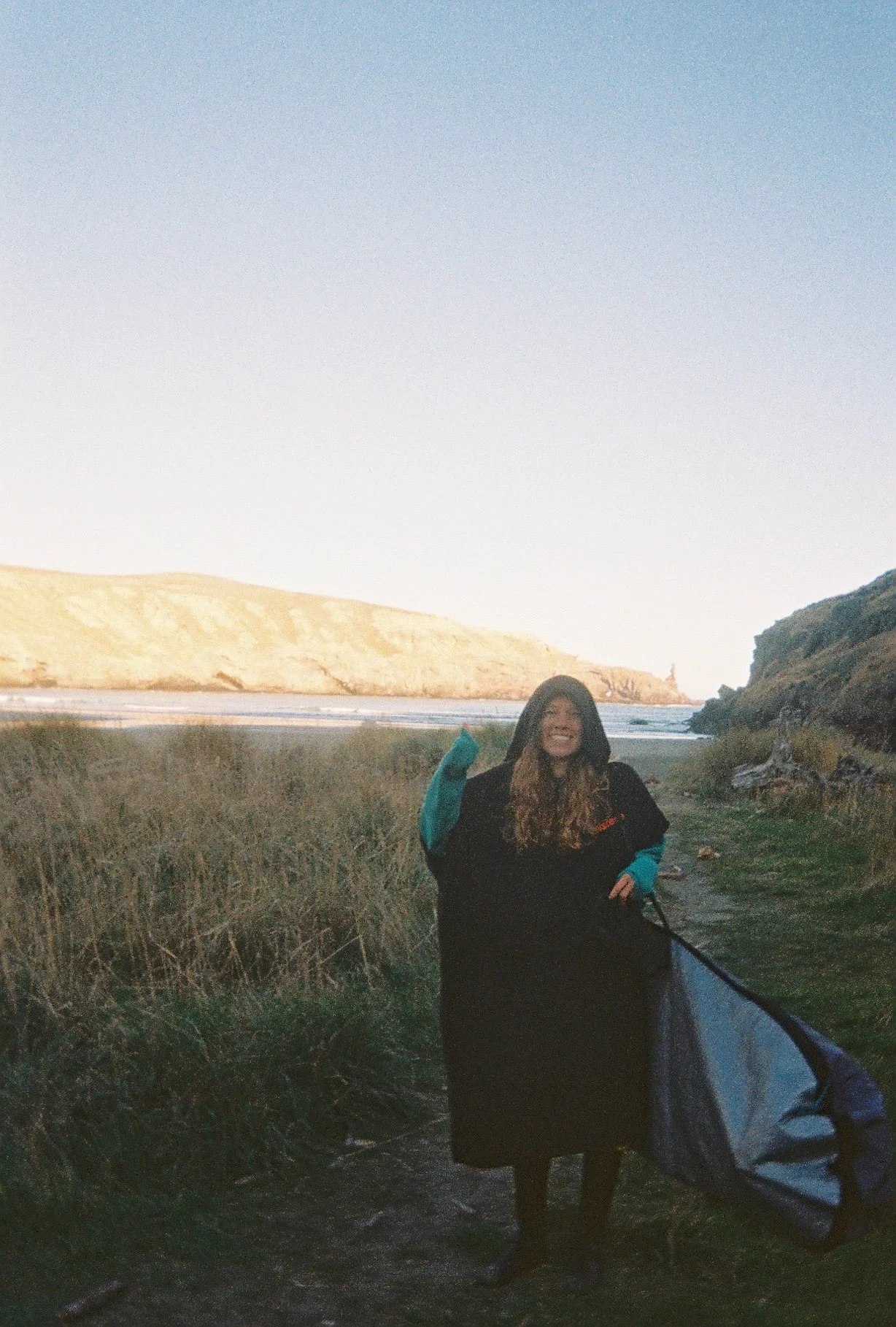 A woman standing outdoors near a coastline with grassy dunes and cliffs, holding a long object, possibly a surfboard or paddle, smiling, during sunset or sunrise.